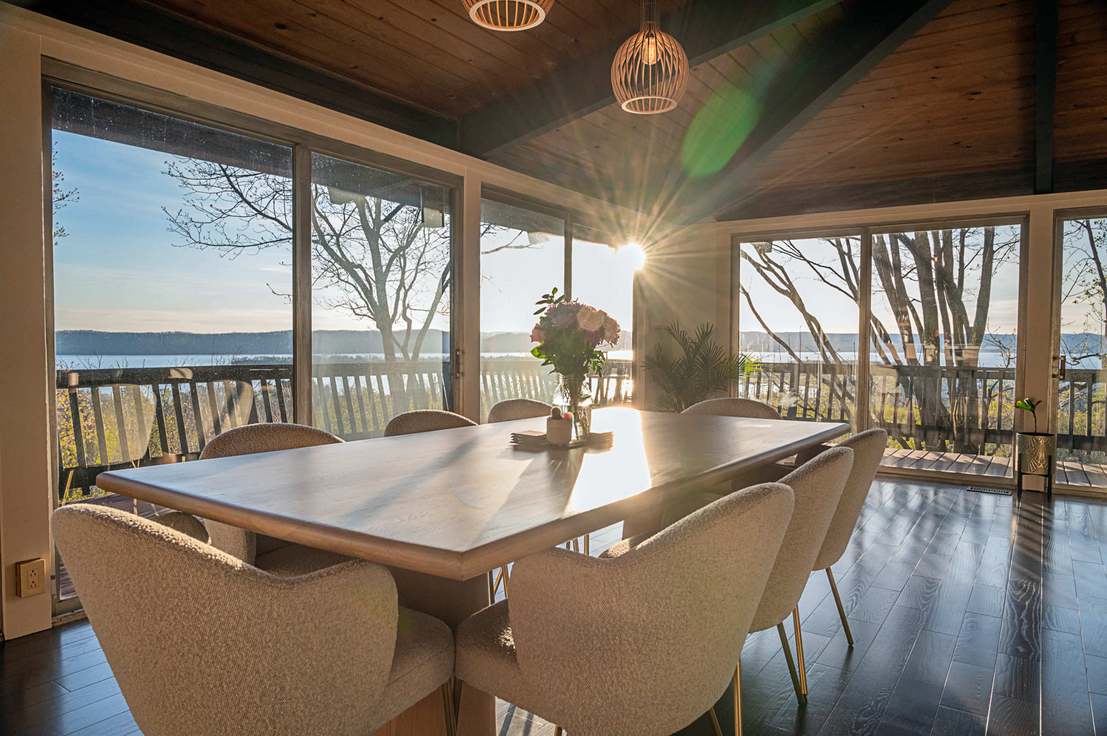 A dining area features a large wooden table surrounded by plush chairs, with sunlight streaming through large windows that overlook a scenic view.