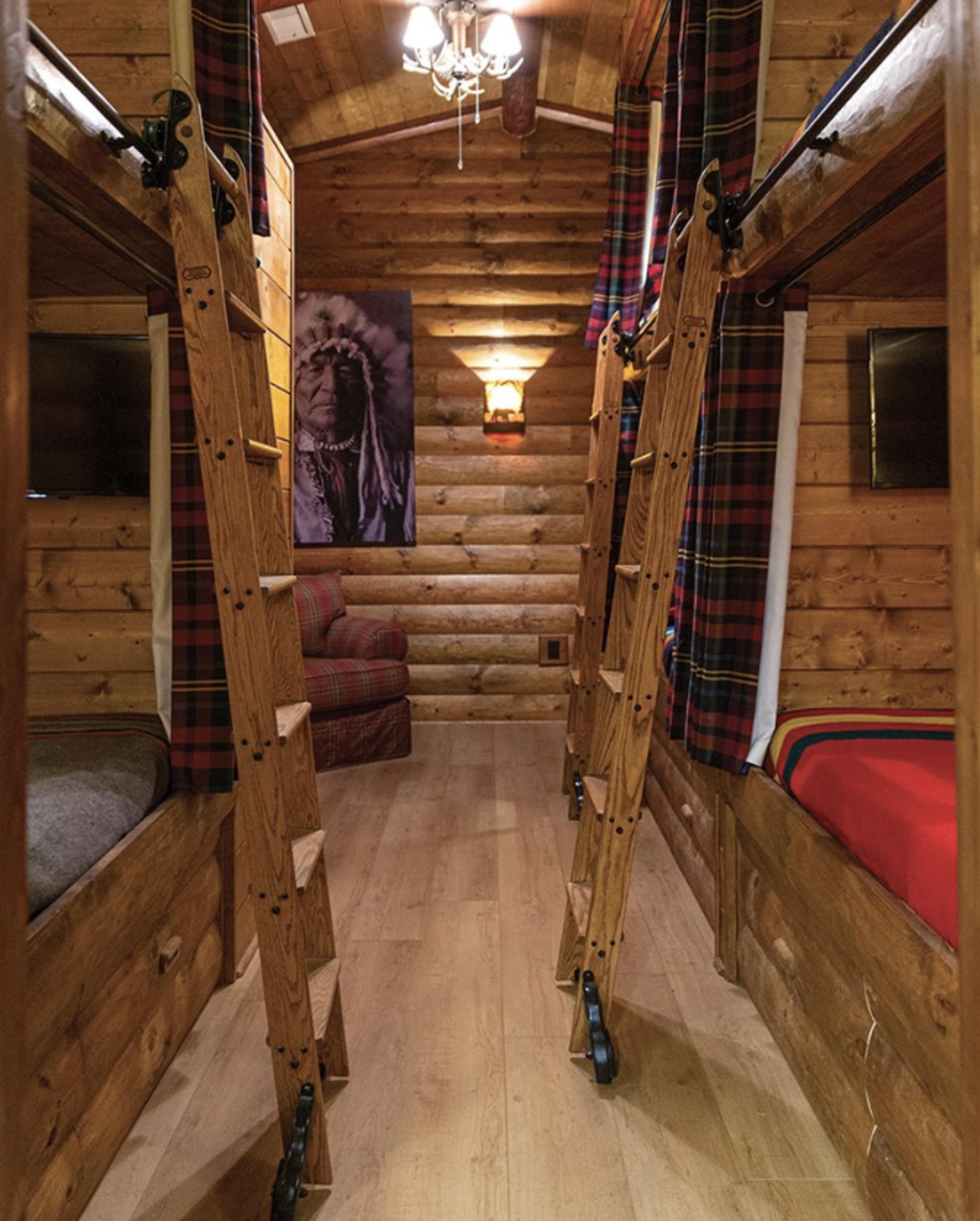 The image features a narrow wooden cabin interior with two sets of bunk beds, each accompanied by wooden ladders, and a cozy seating area at the end.