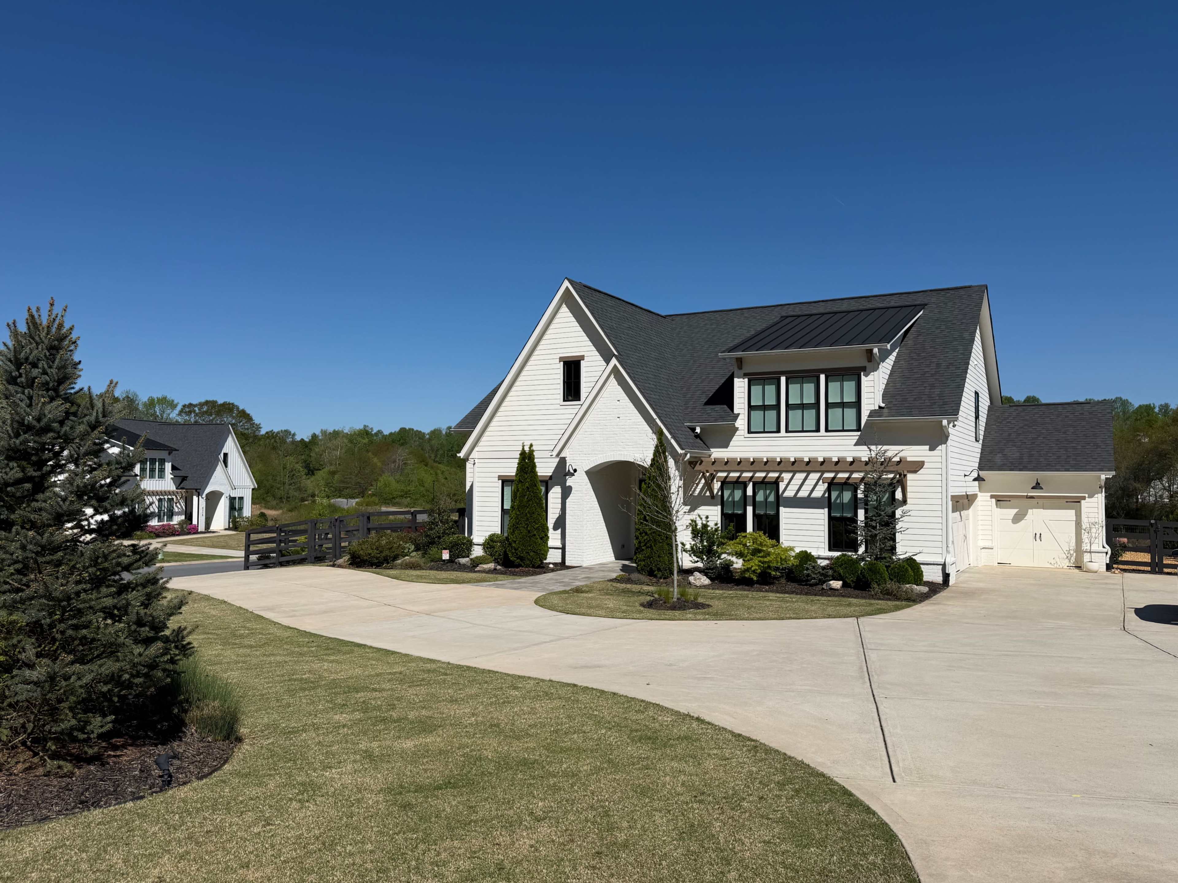 A modern white farmhouse features a landscaped yard with a circular driveway and a clear blue sky overhead.