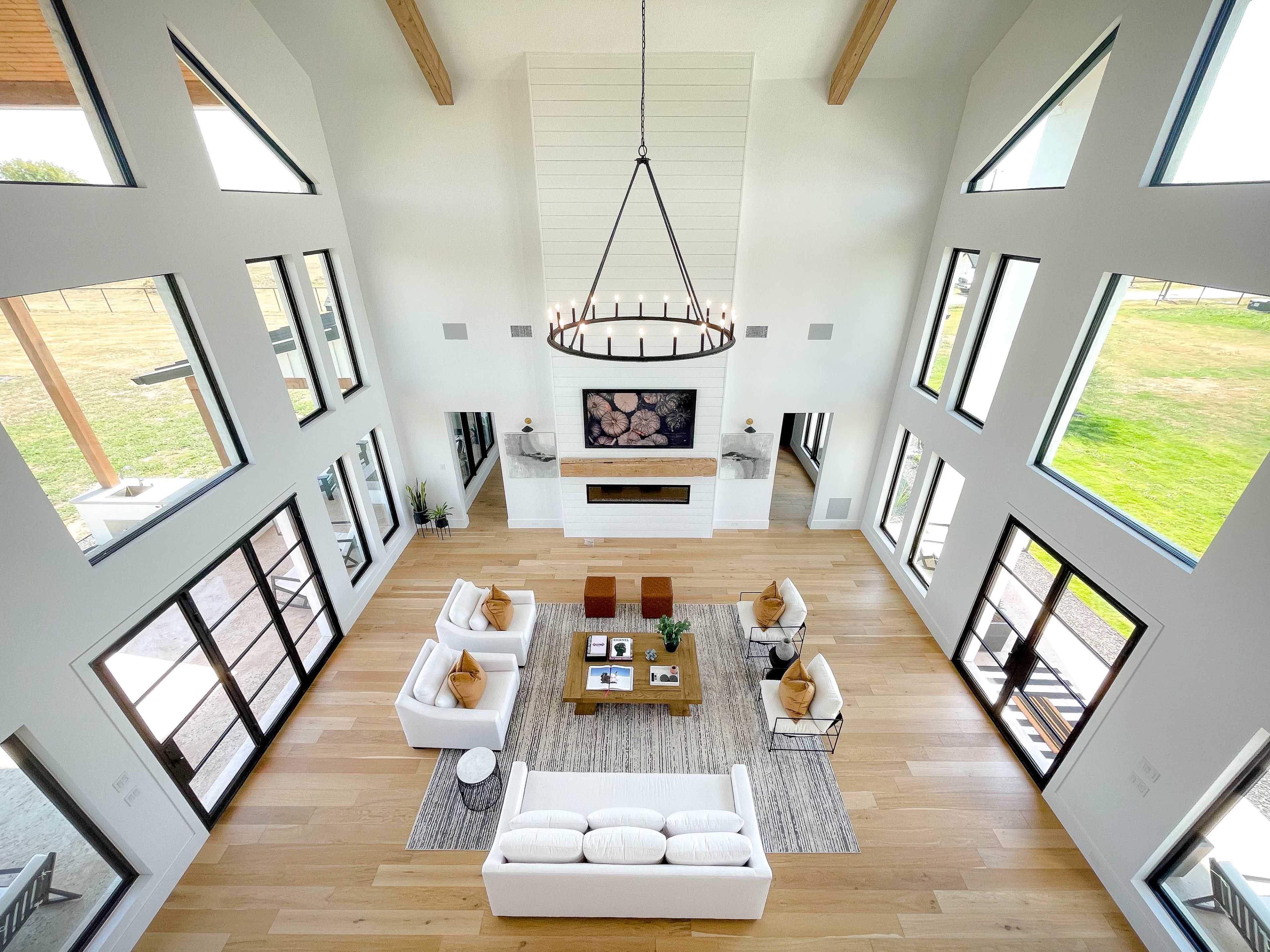 The image shows a spacious, modern living room with large windows, high ceilings, and a central seating area arranged around a coffee table.