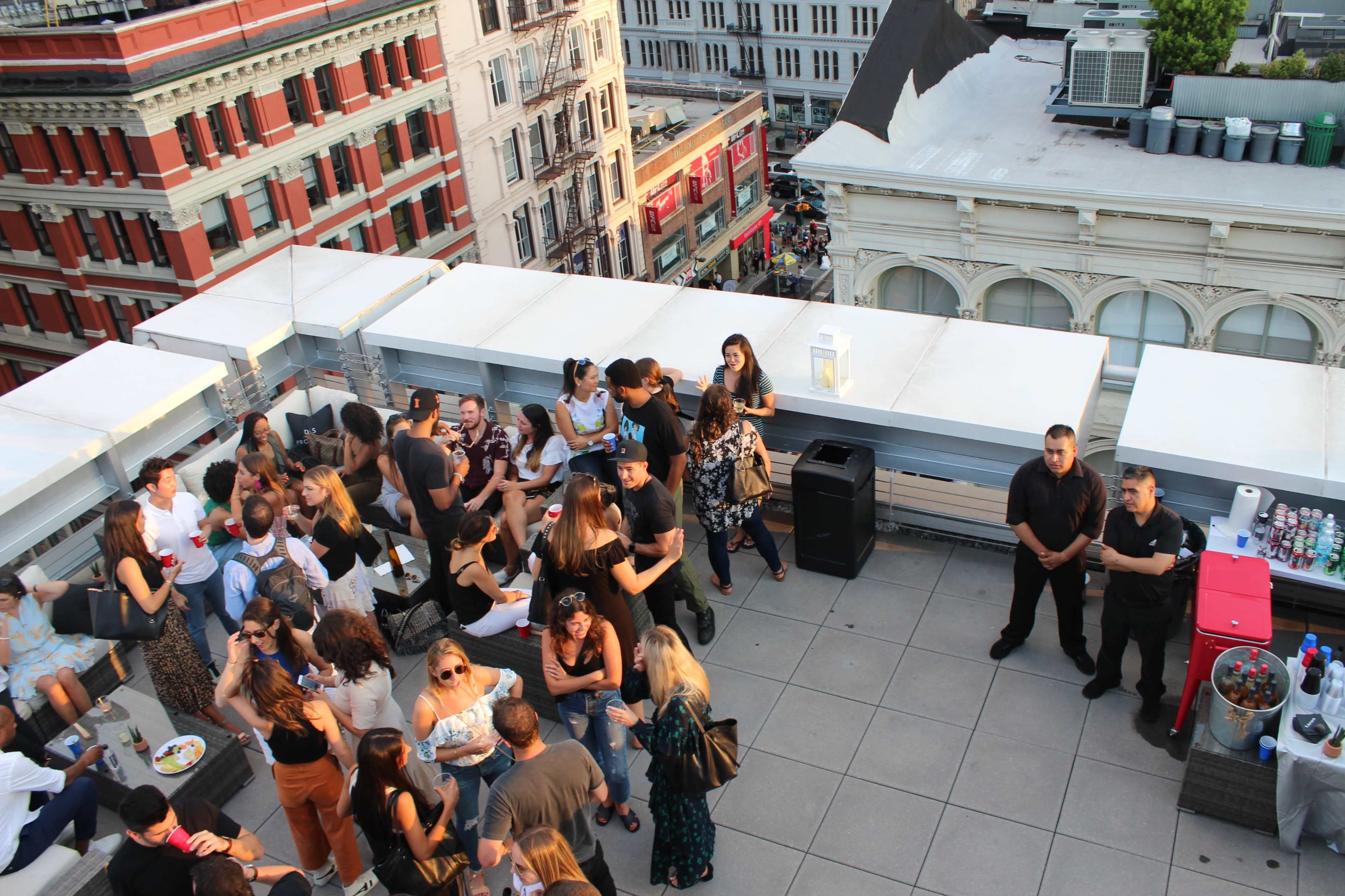 A large group of people socializes on a rooftop terrace with views of city buildings and vehicles in the background.