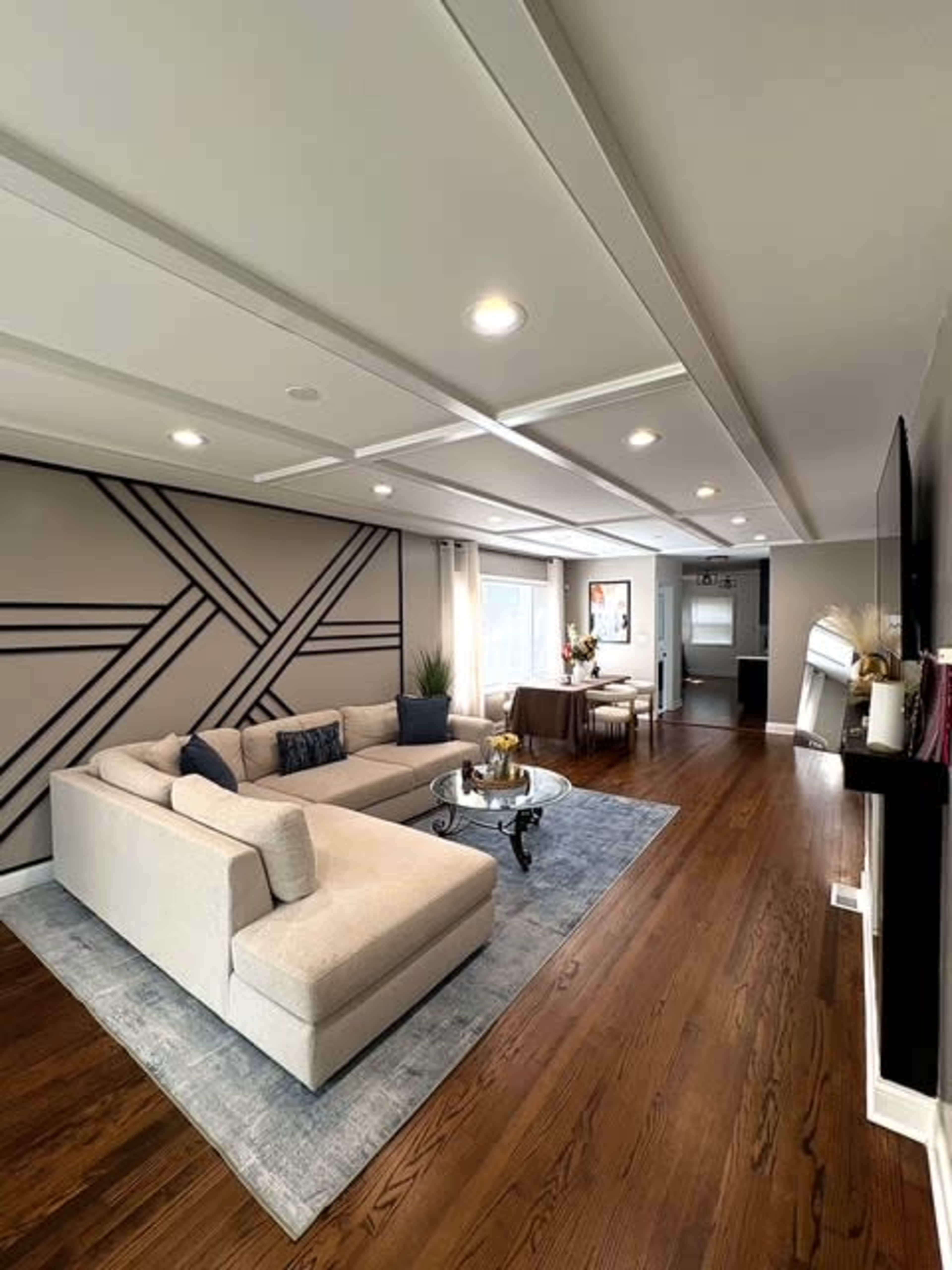 The living room features a spacious sectional sofa, a decorative area rug, and modern lighting, with a patterned wall and large windows allowing natural light to enter.