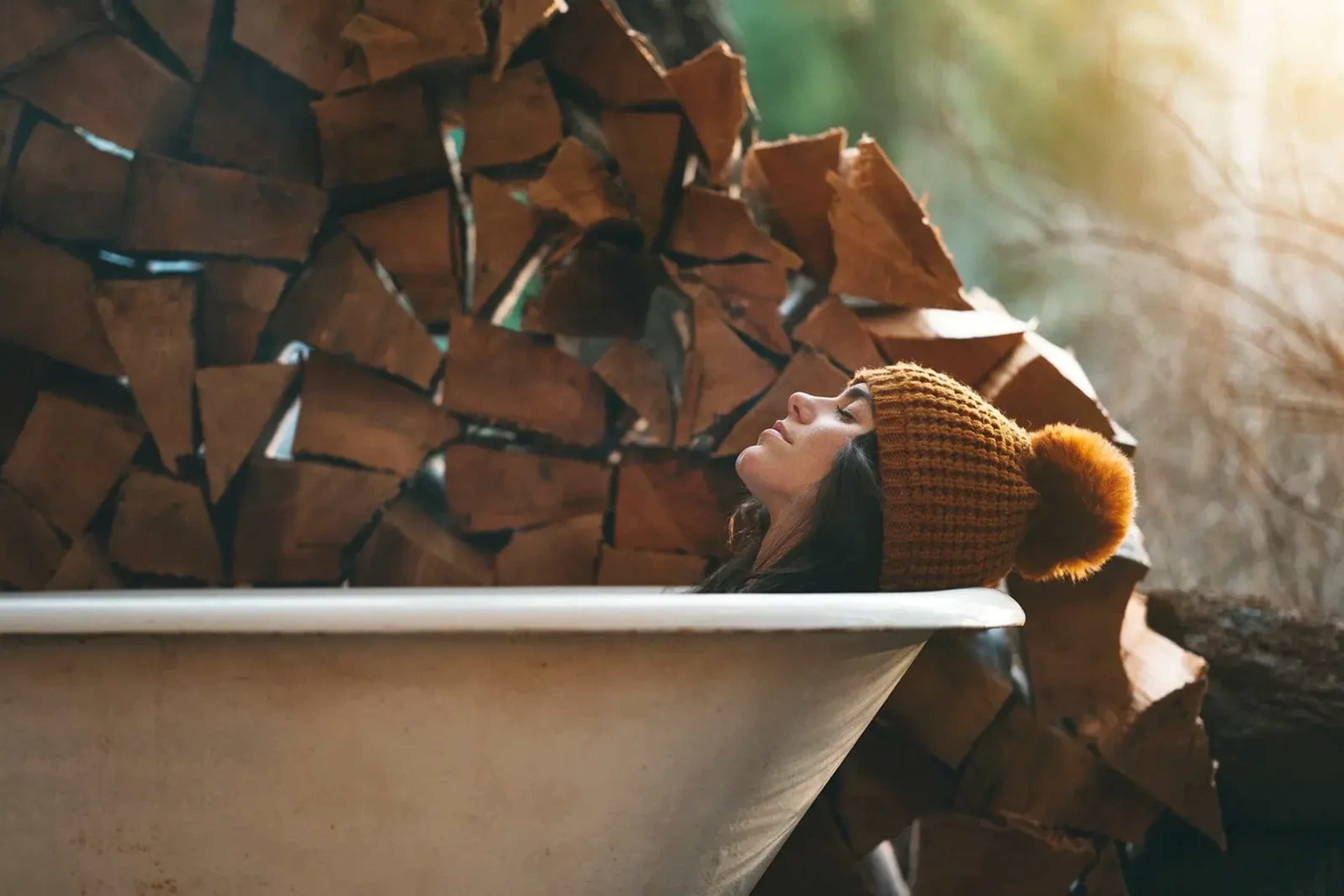 A woman wearing a knitted hat relaxes in a vintage bathtub beside a stack of firewood.