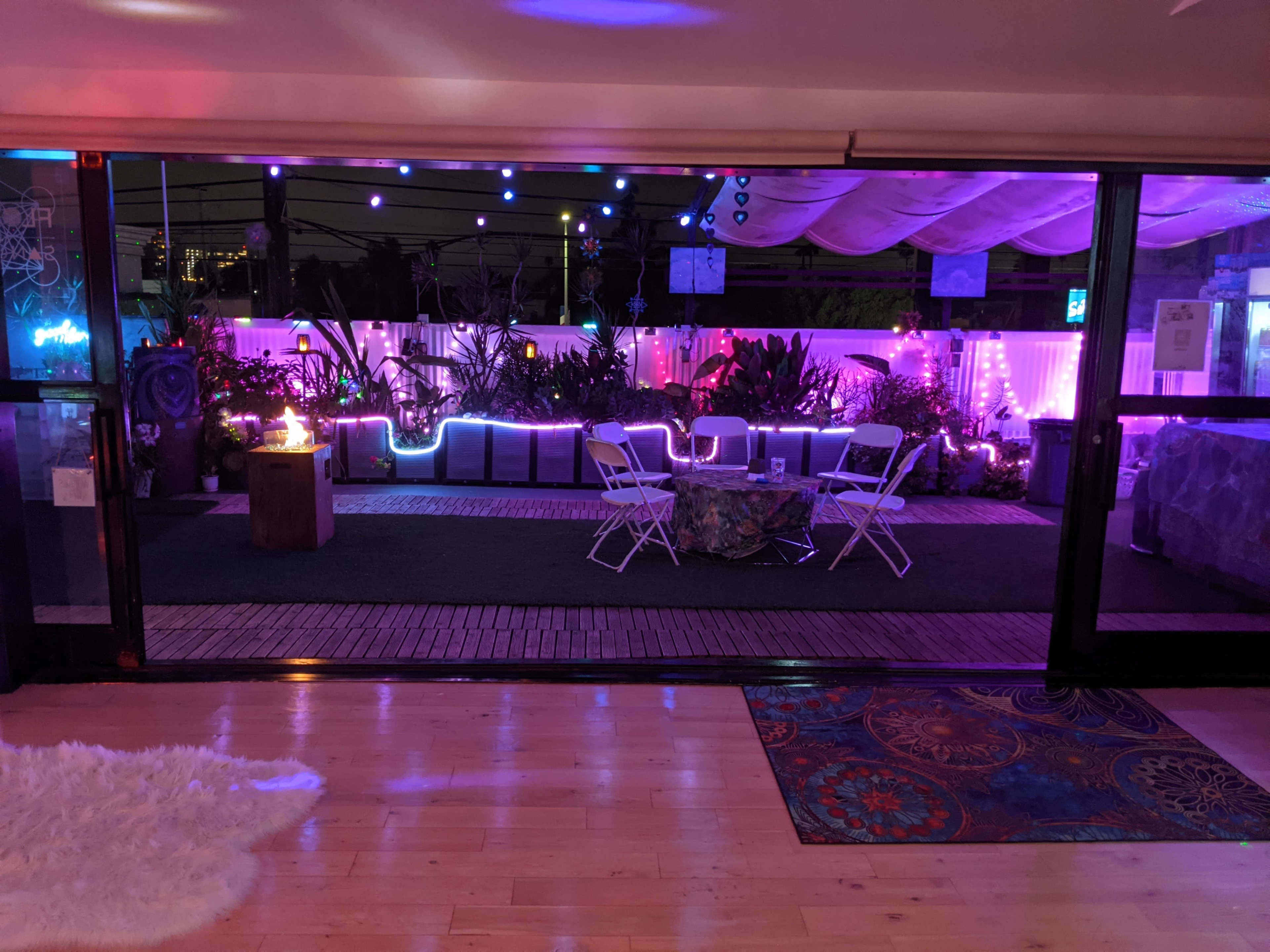 A well-lit outdoor lounge area features colorful lighting, seating arrangements, and decorative plants visible through a large glass doorway.