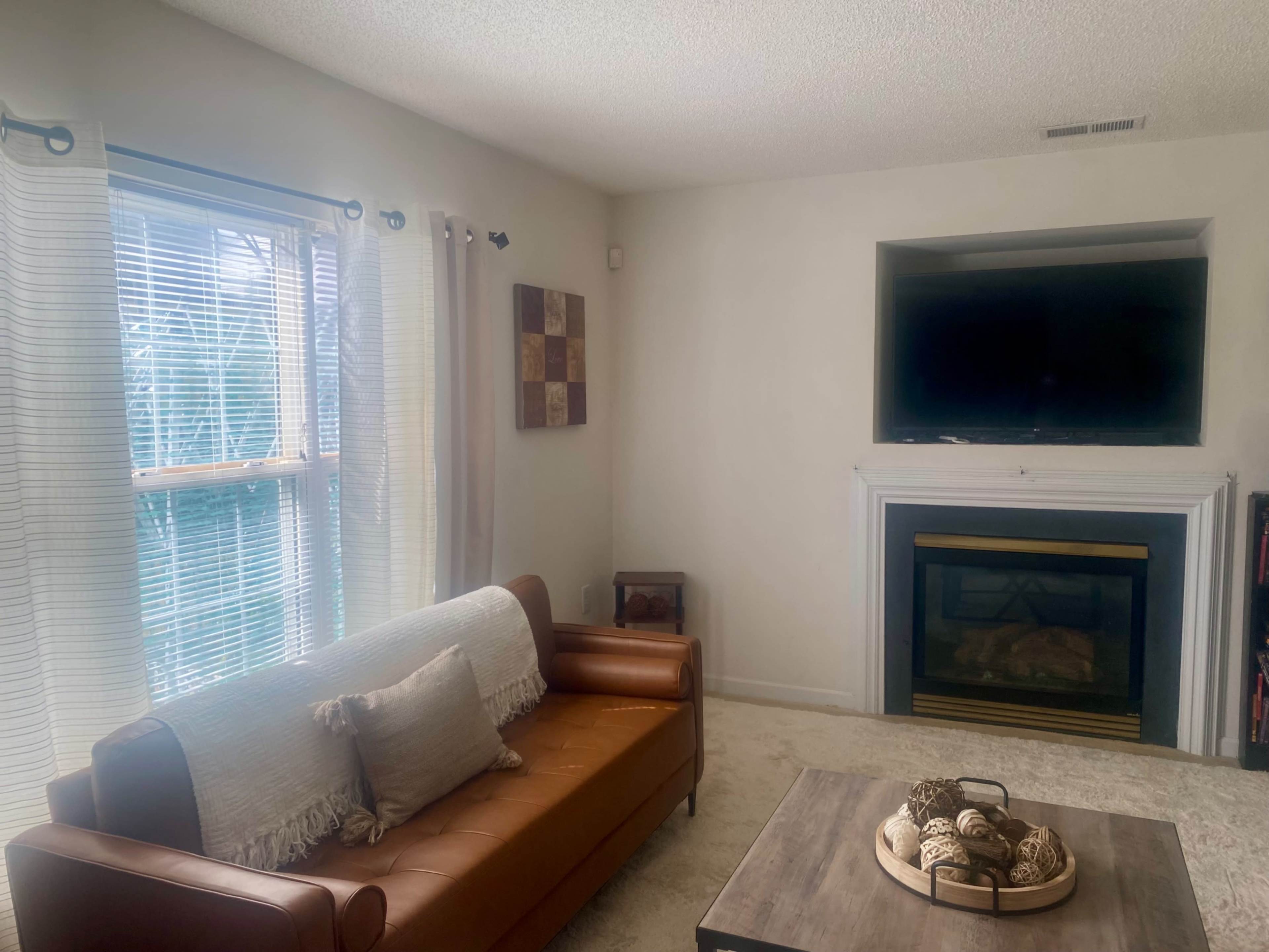 A living room features a brown leather couch, a wooden coffee table with decorative items, and a wall-mounted television above a fireplace.