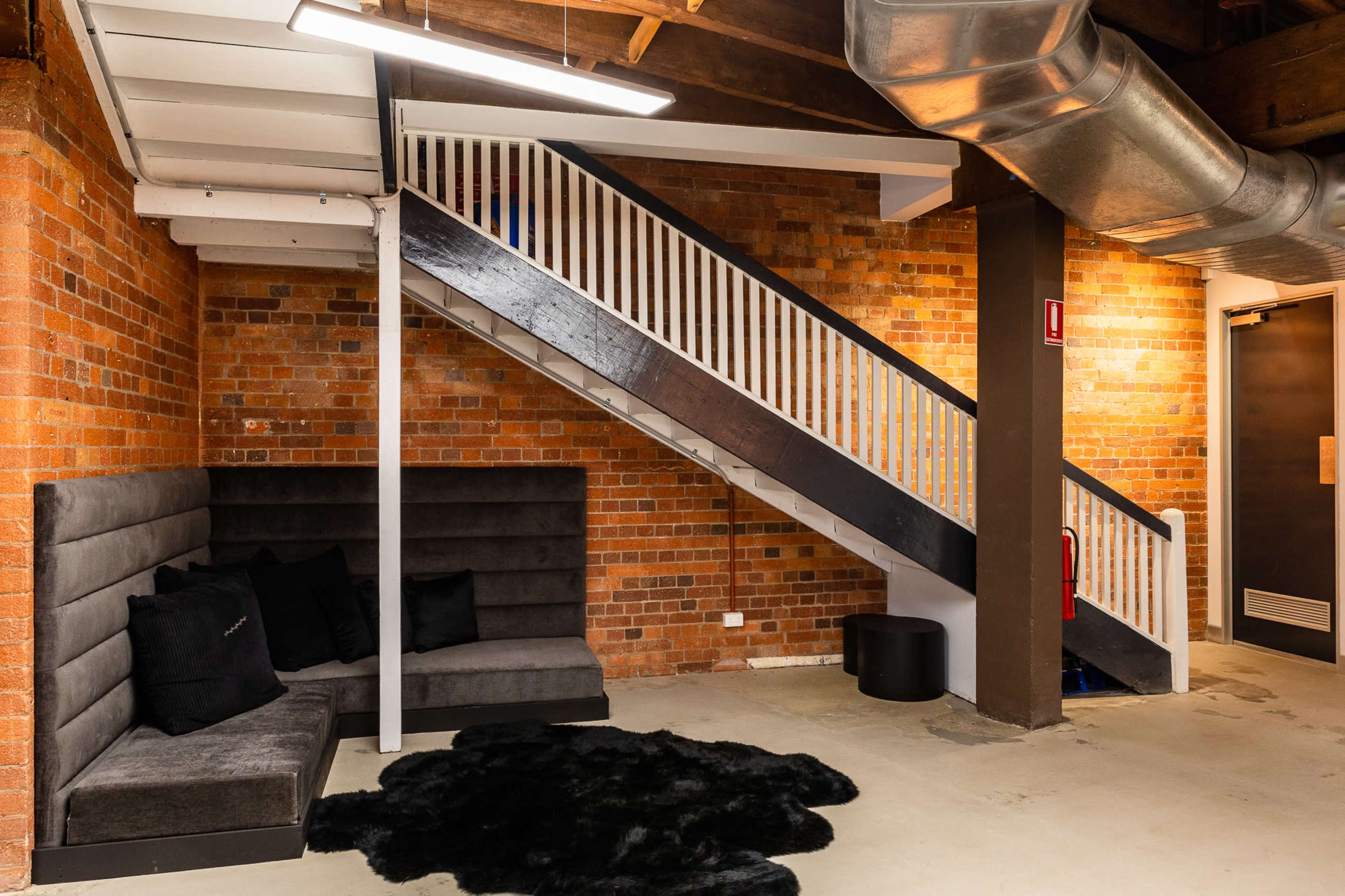 The image shows a minimalist lounge area featuring a black upholstered seating arrangement next to a staircase with a wooden and metal railing in a brick-walled basement space.