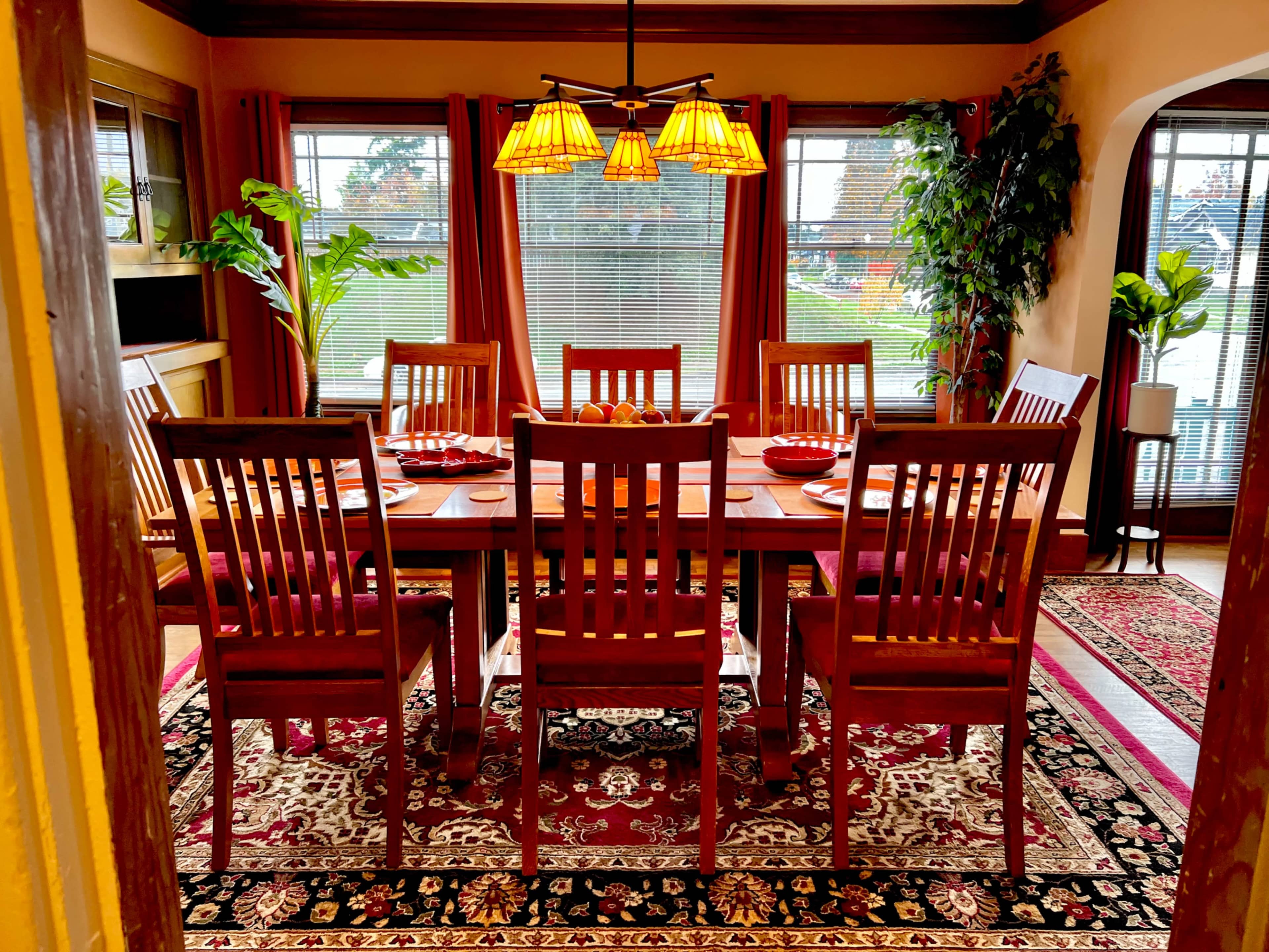 A dining room features a wooden table set for eight, surrounded by matching chairs, with decorative plants and large windows allowing natural light.