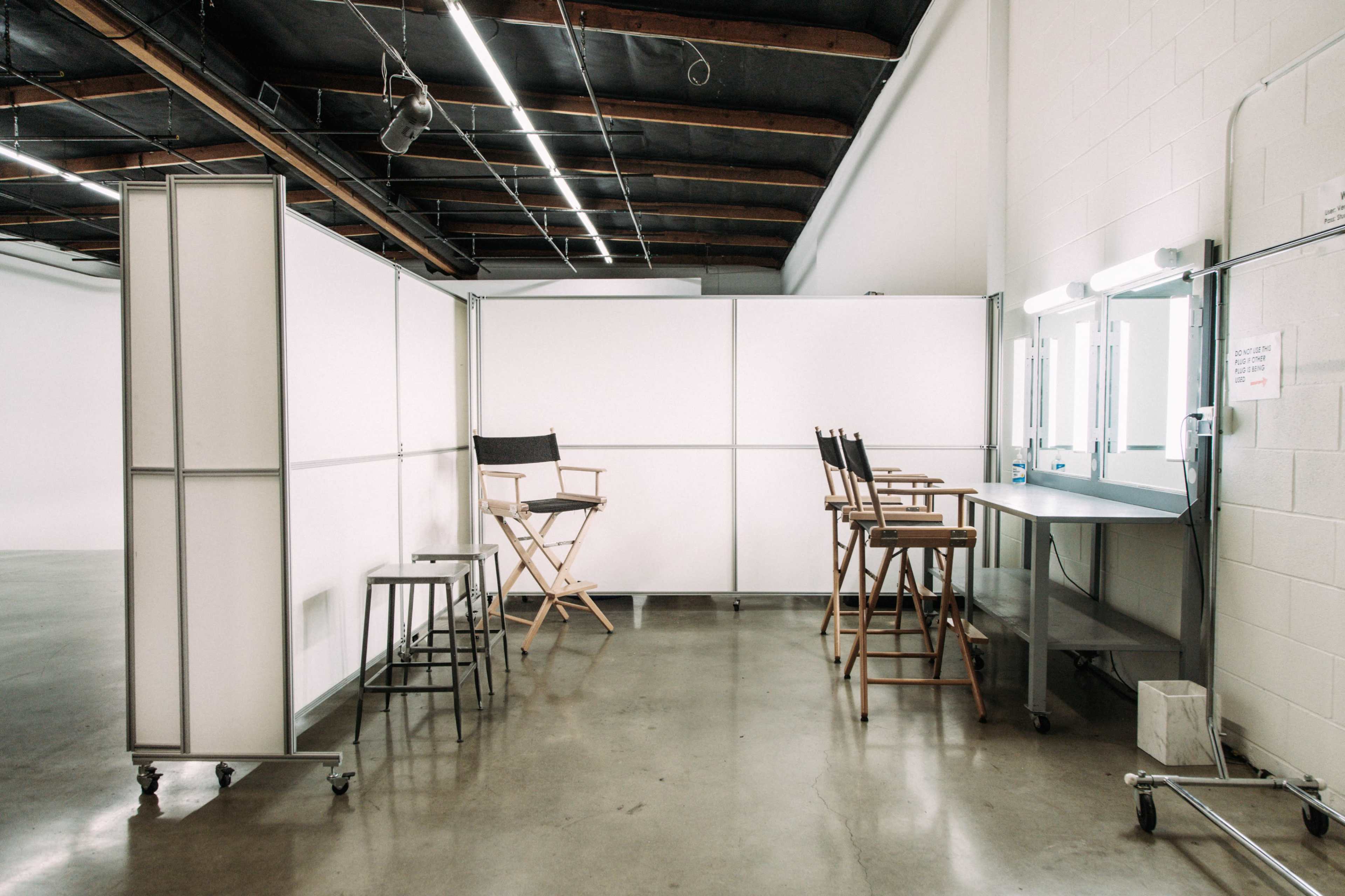 A clean, well-lit makeup and styling area featuring several mirrored stations and chairs separated by white partitions.