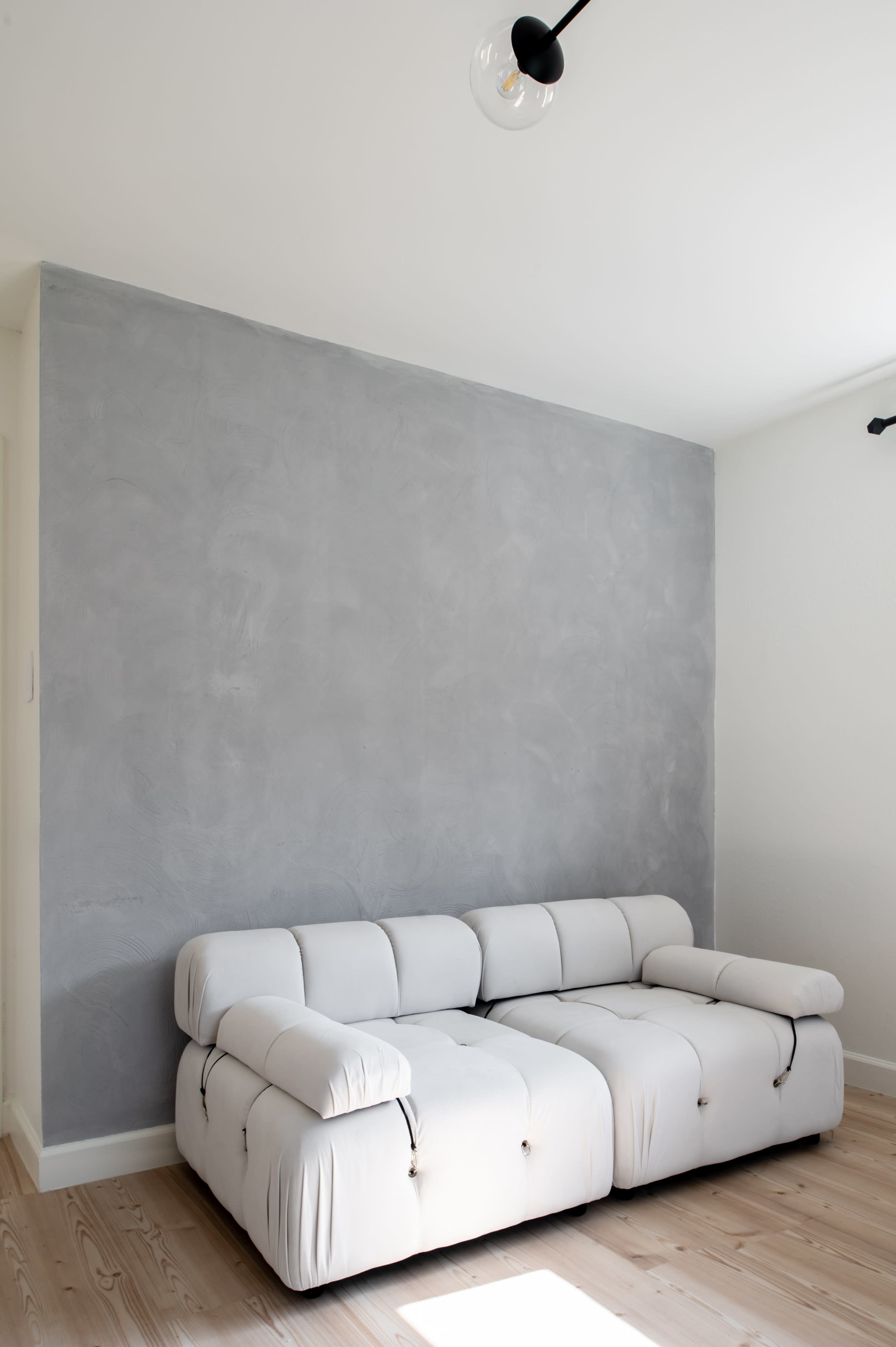 A white sectional sofa is positioned against a light gray textured wall in a minimalistic room with wooden flooring.
