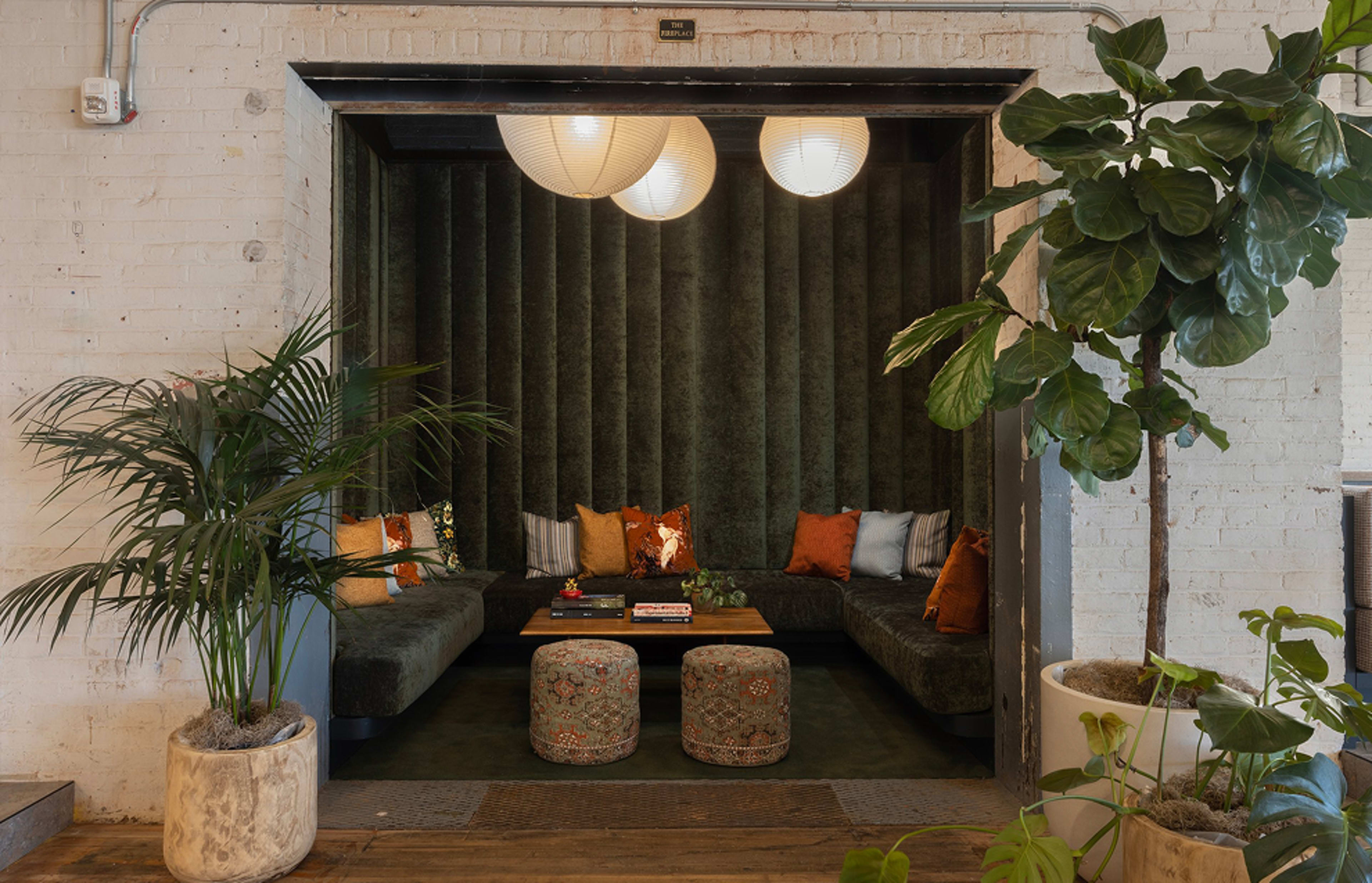 The image shows a cozy seating area with plush green upholstery, decorative pillows, a wooden coffee table, and two patterned stools, framed by plants and illuminated by hanging lanterns.
