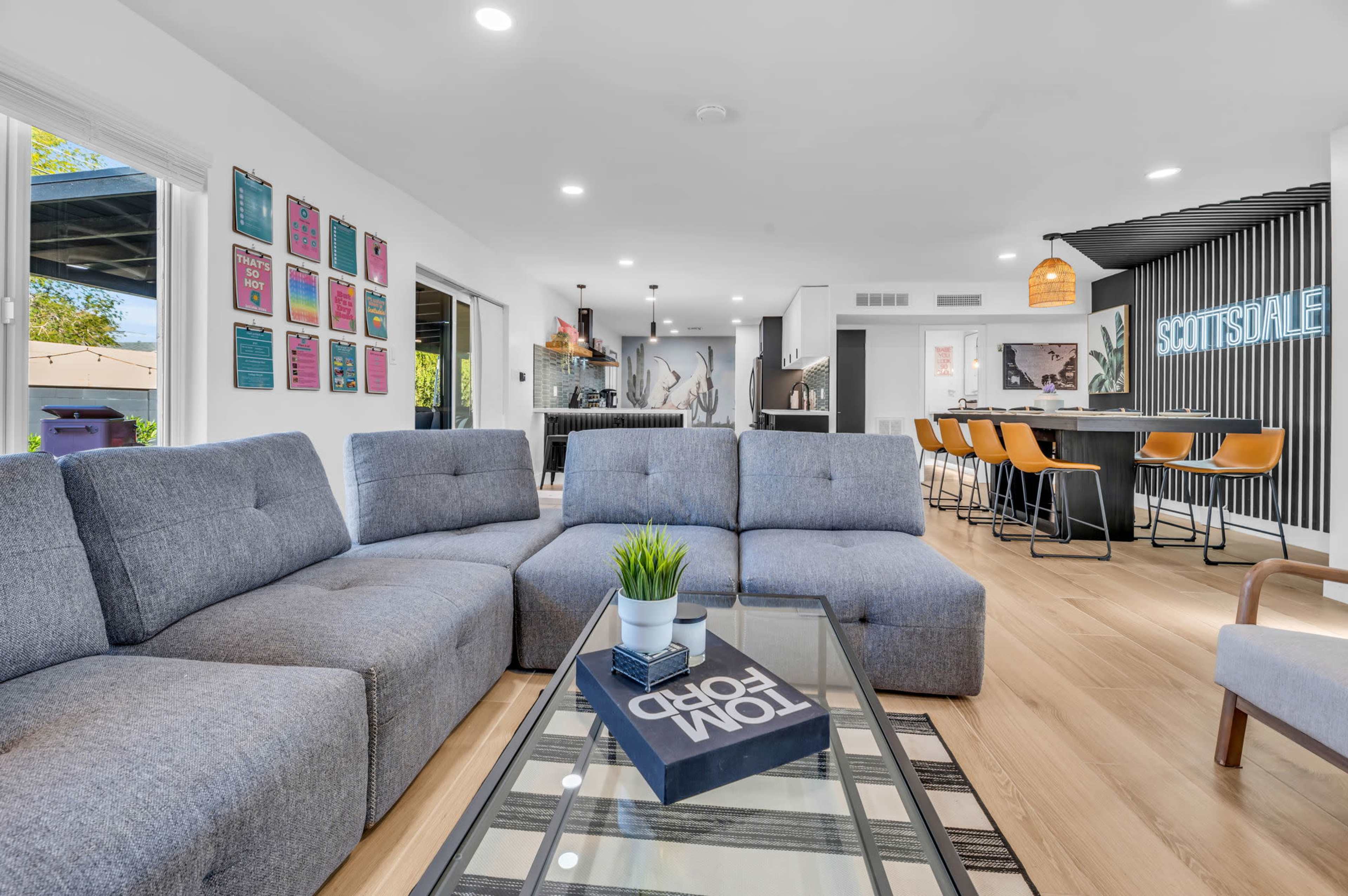 A modern living room features a gray sectional sofa, a glass coffee table, and a contemporary kitchen bar with orange stools in the background.