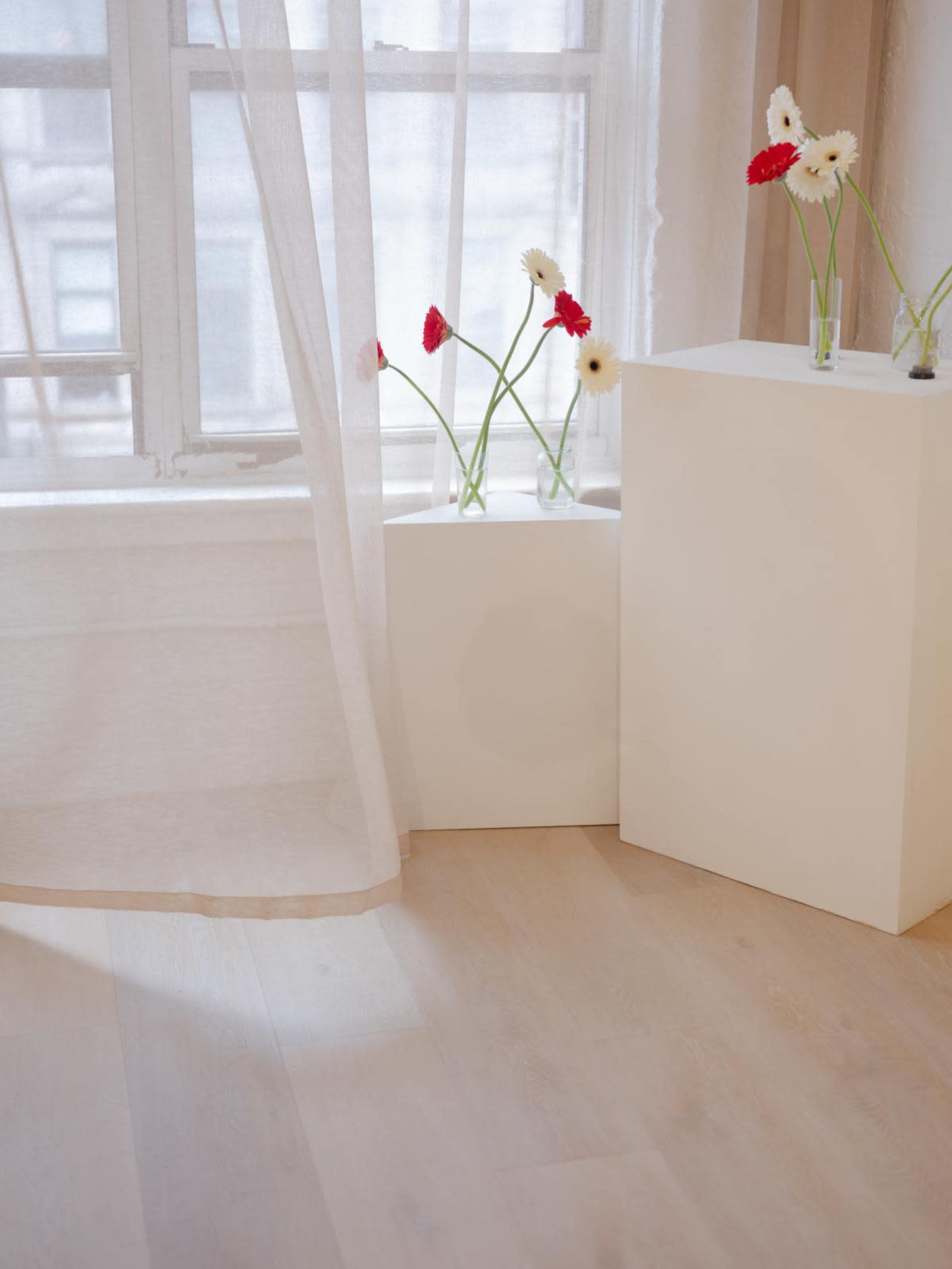 The image shows a corner of a room with light wooden flooring, a sheer curtain, and two small white pedestals holding vases with flowers.