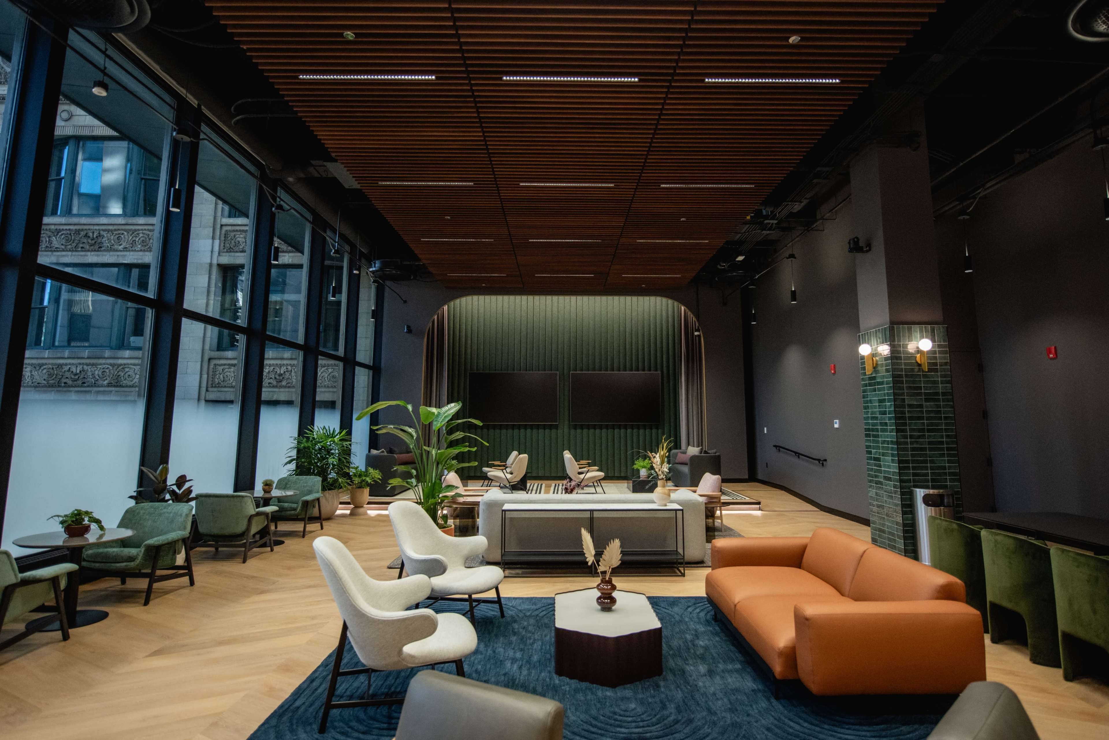 The image shows a modern lounge area with a mix of seating, including armchairs and sofas, large windows, and wooden ceiling panels.