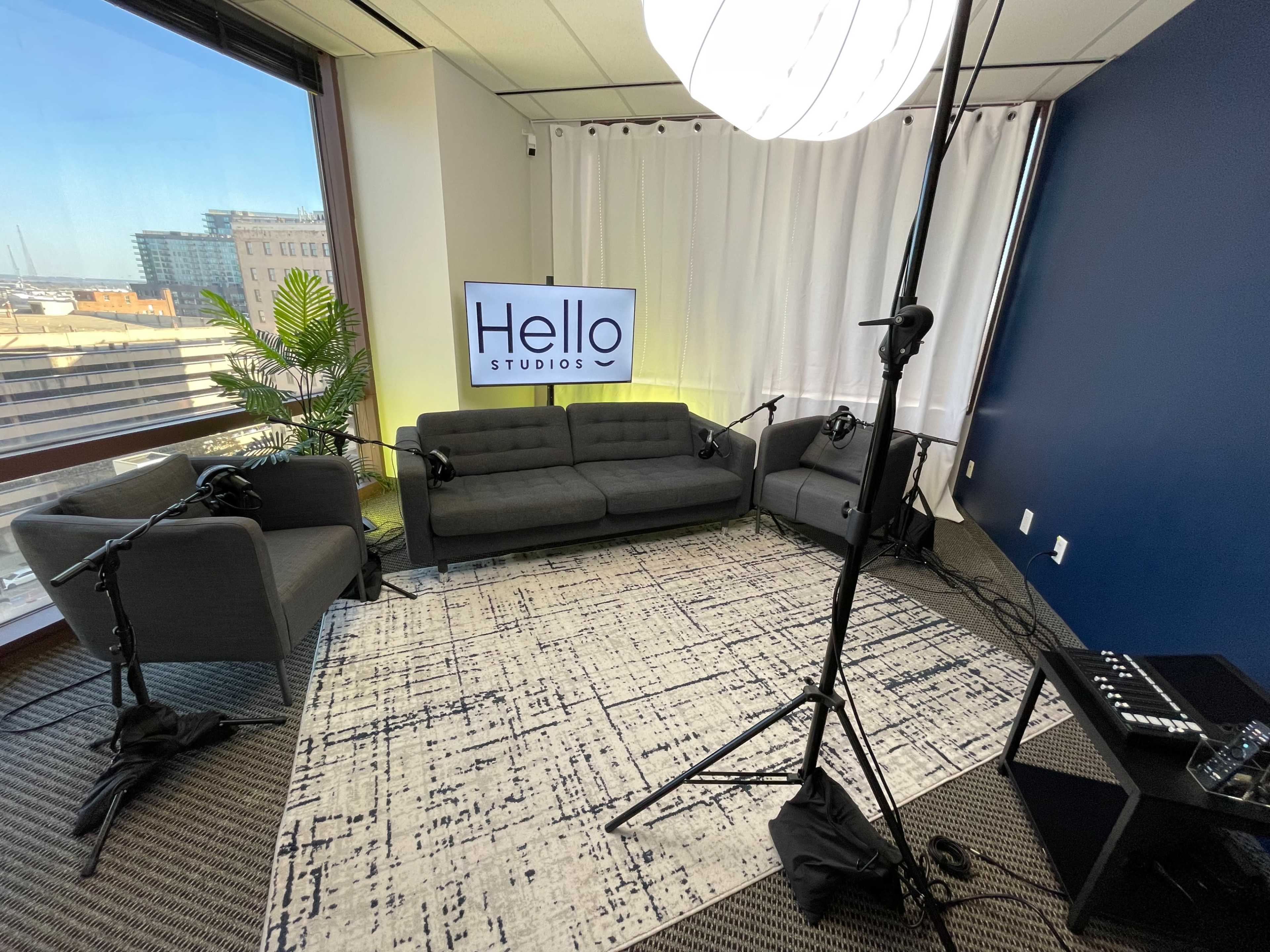 A modern studio is set up with a gray sofa, two microphones on stands, and a television screen displaying "Hello Studios," complemented by a large window offering a city view.