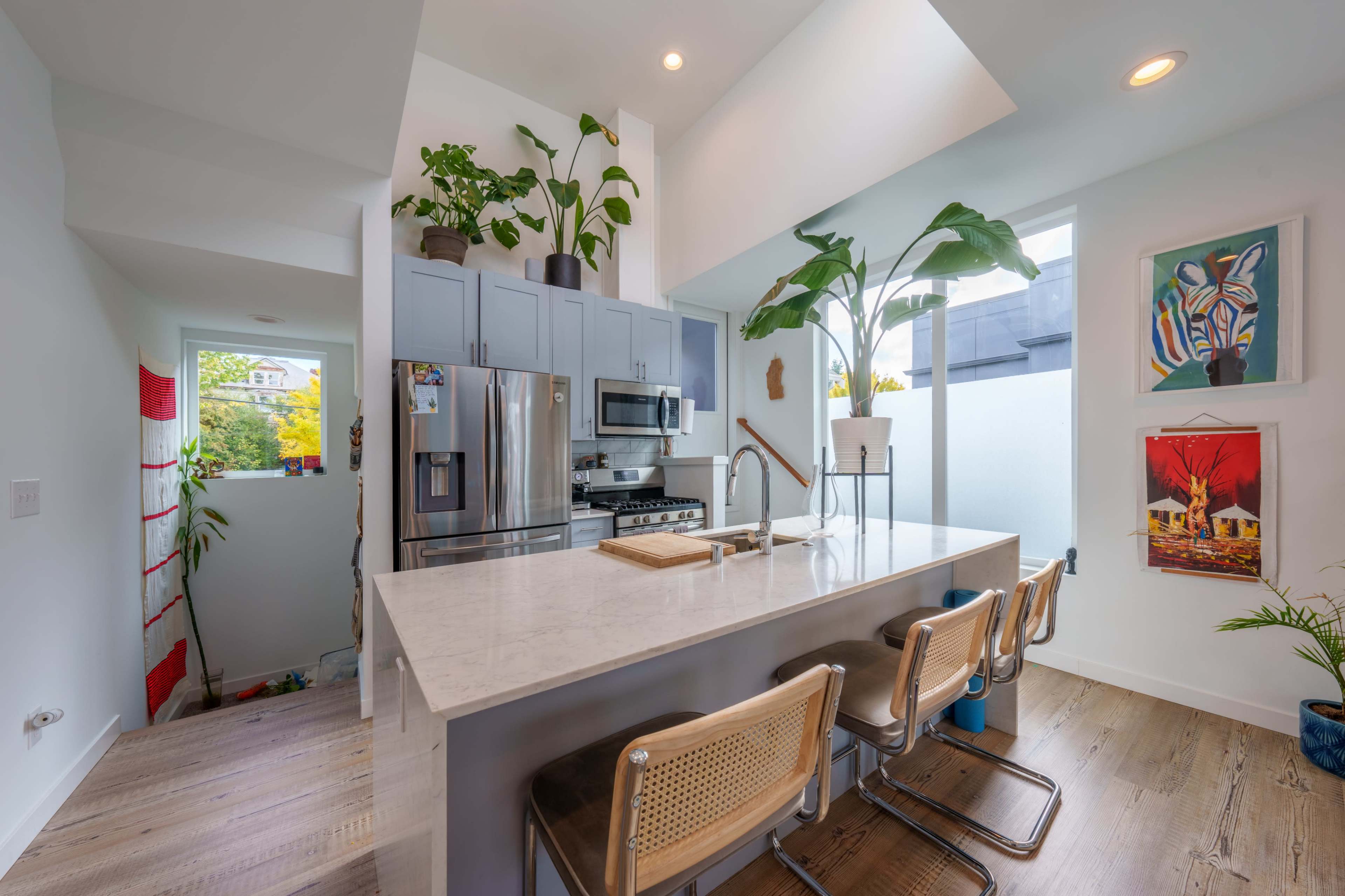 A modern kitchen features a large island with bar stools, stainless steel appliances, and abundant plants on the countertop and shelves.