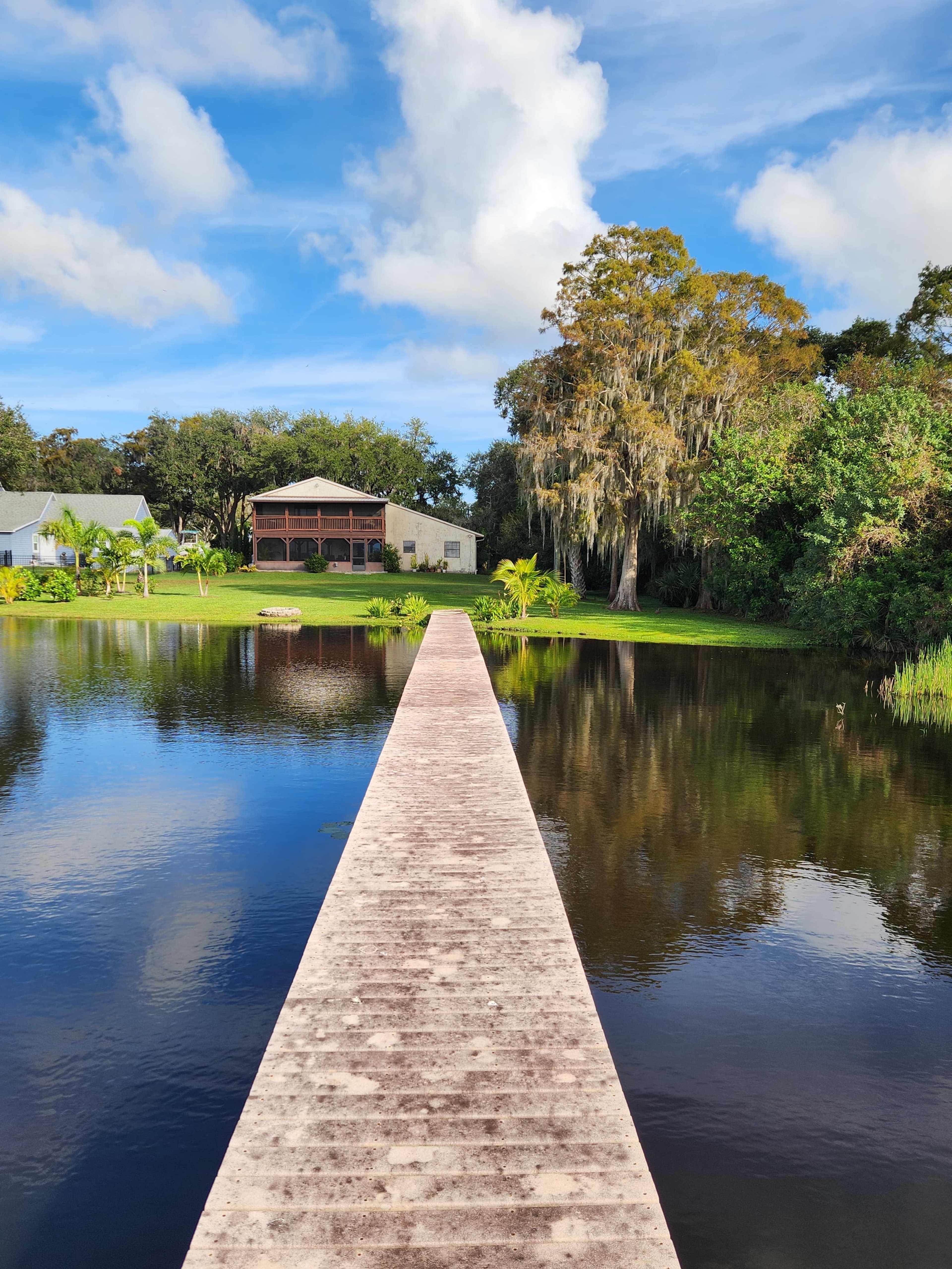 A concrete pathway extends into a calm lake, flanked by trees and buildings on either side.