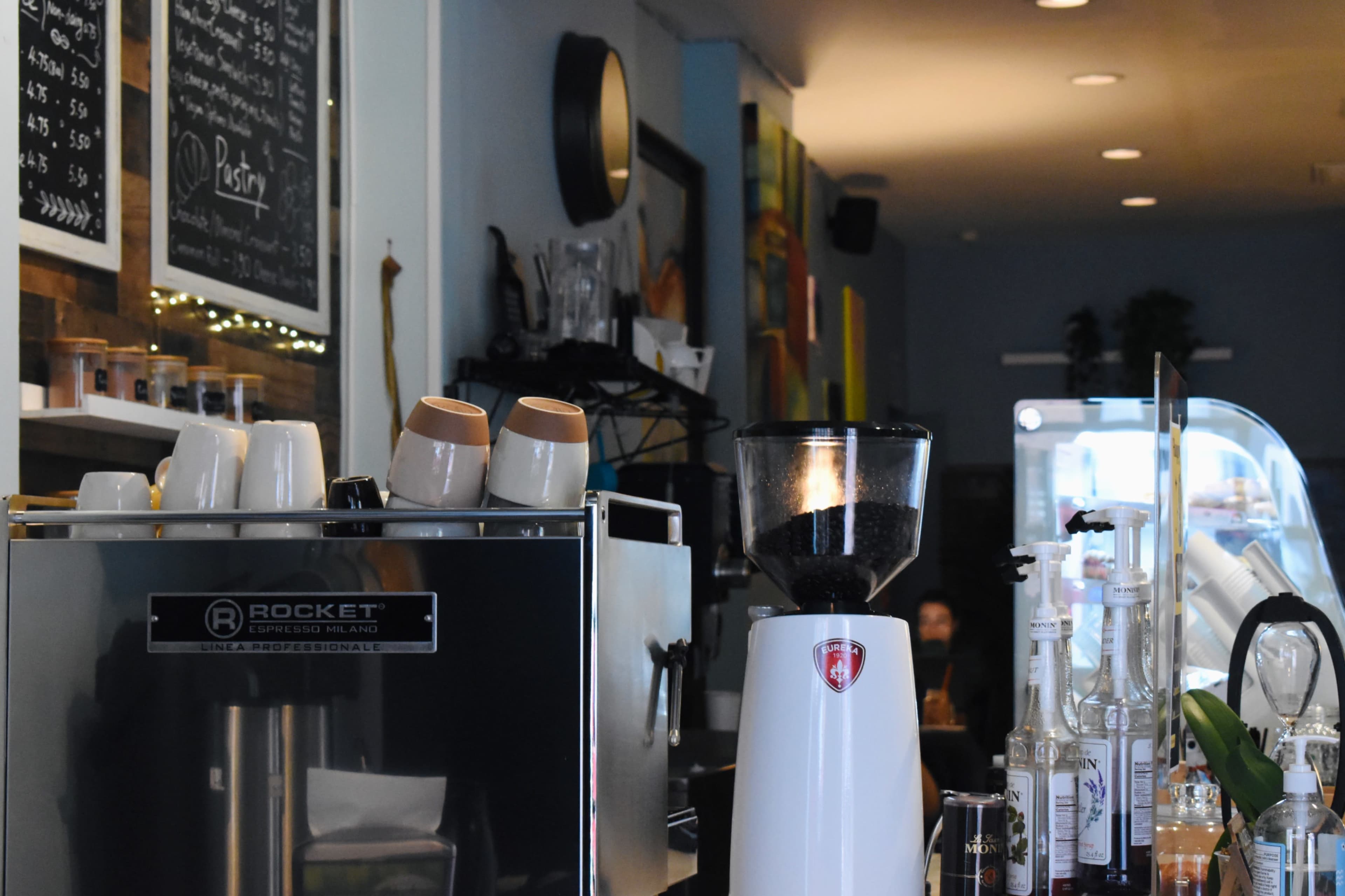The image features a coffee shop interior with an espresso machine, a coffee grinder, and neatly stacked cups on a counter, surrounded by shelves displaying menu items and pastries.
