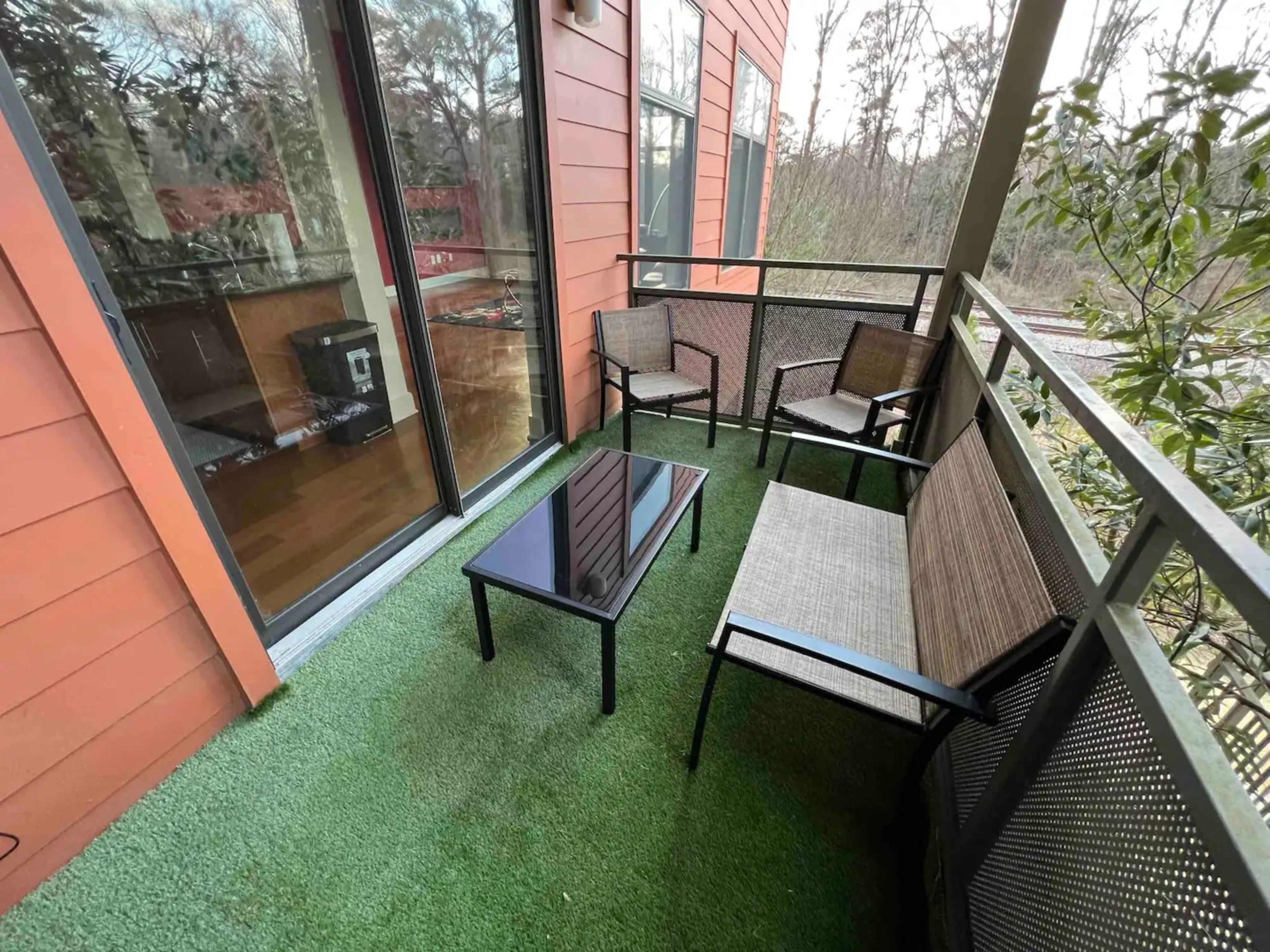 A balcony features a glass-top table surrounded by four chairs on artificial grass with a view of trees in the background.