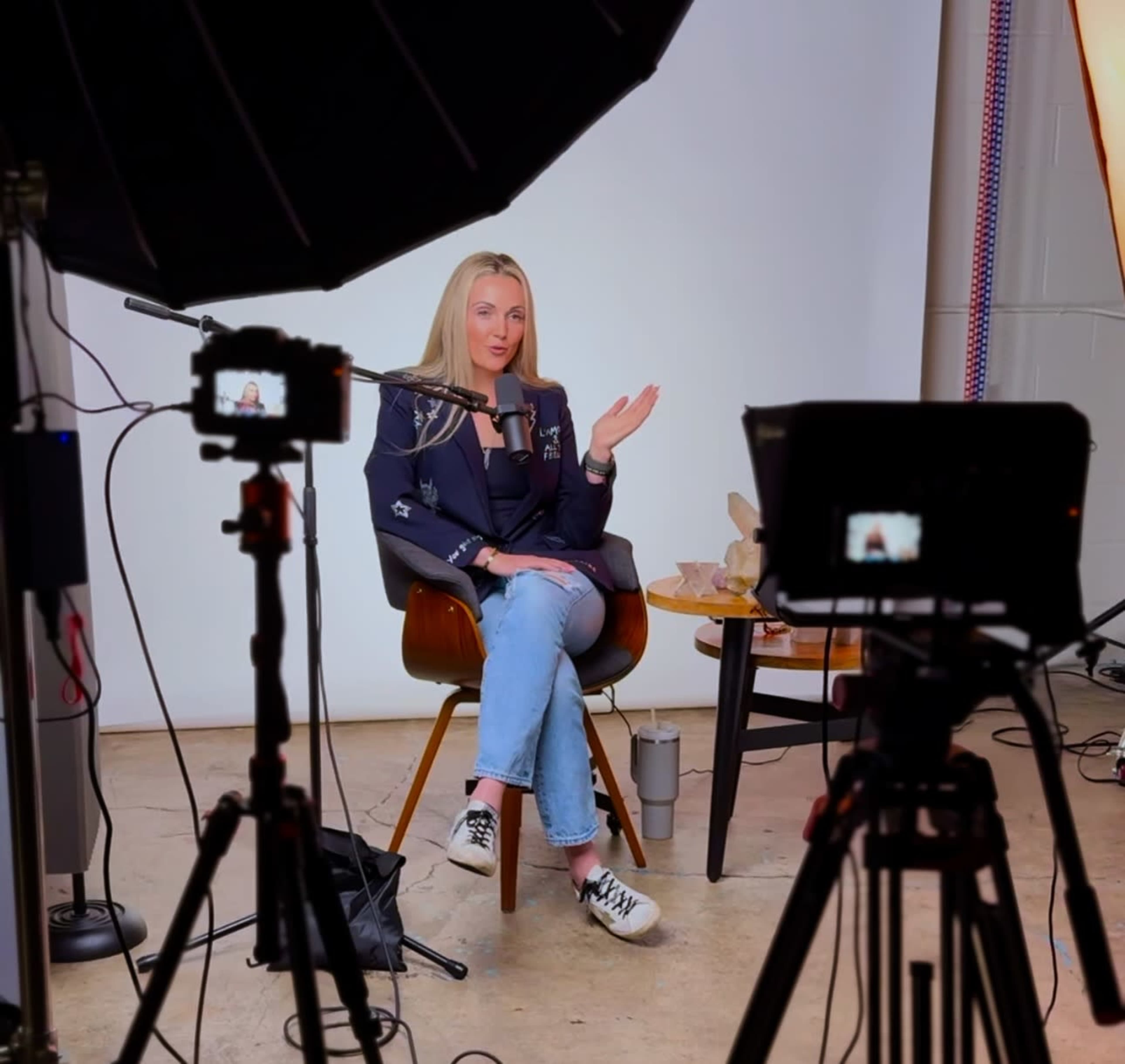 A person sits in a chair in front of a microphone, surrounded by cameras and studio lighting, in a recording setup.