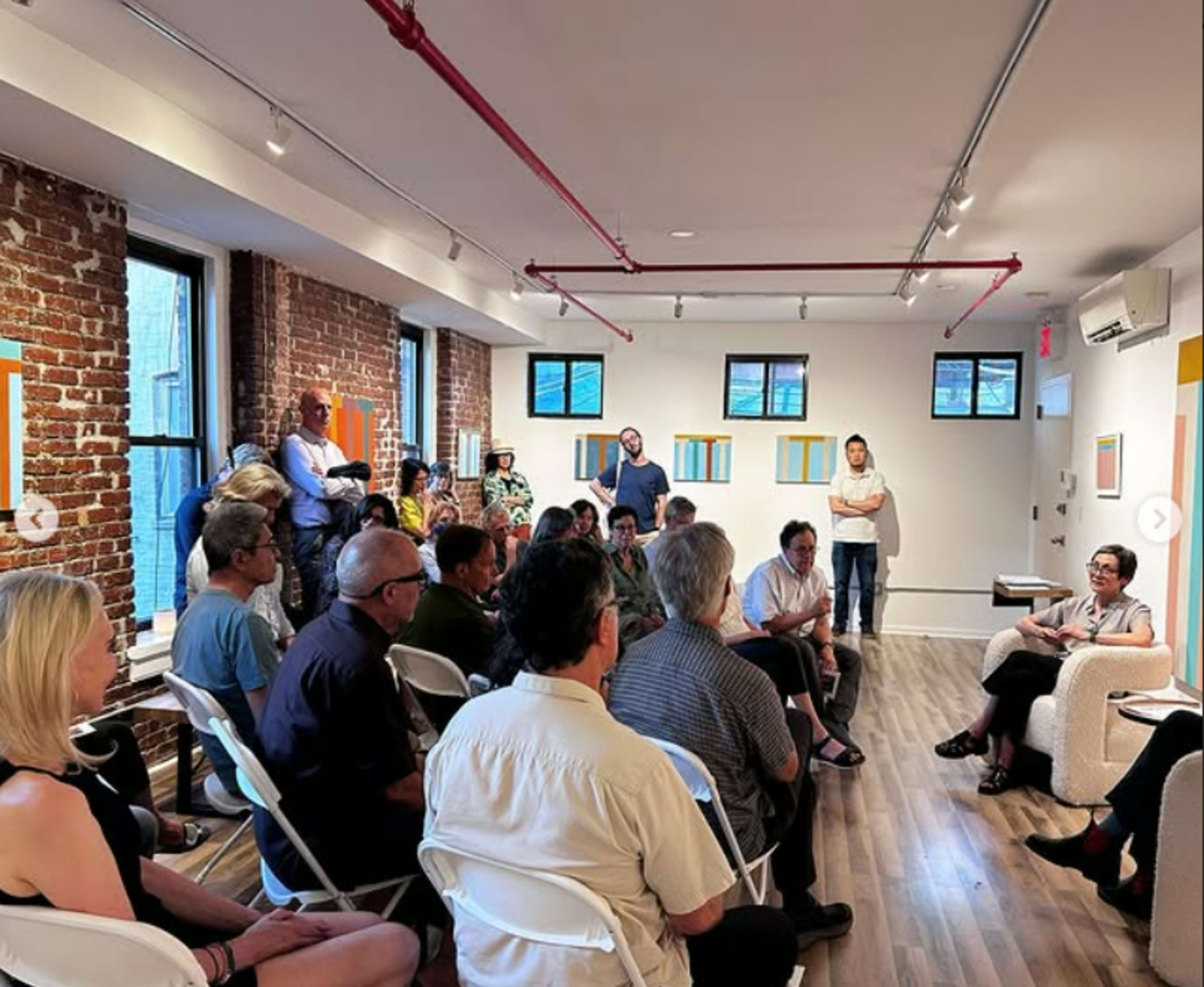 A group of people sits in a modern art gallery, engaging in a discussion while surrounded by abstract artwork on the walls.