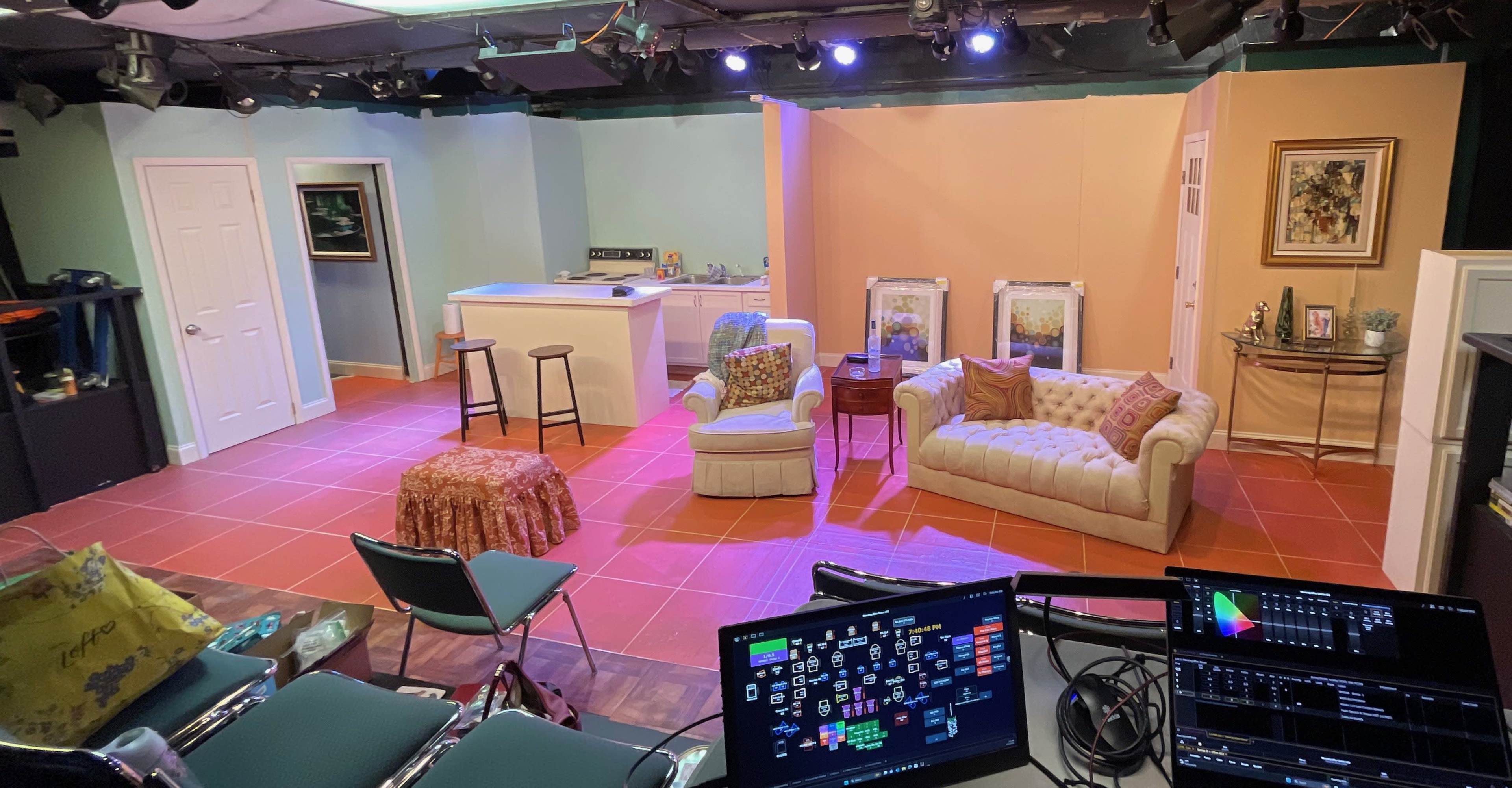 A small stage set is arranged with a kitchen area, two seating areas, and decorative elements in a theater.