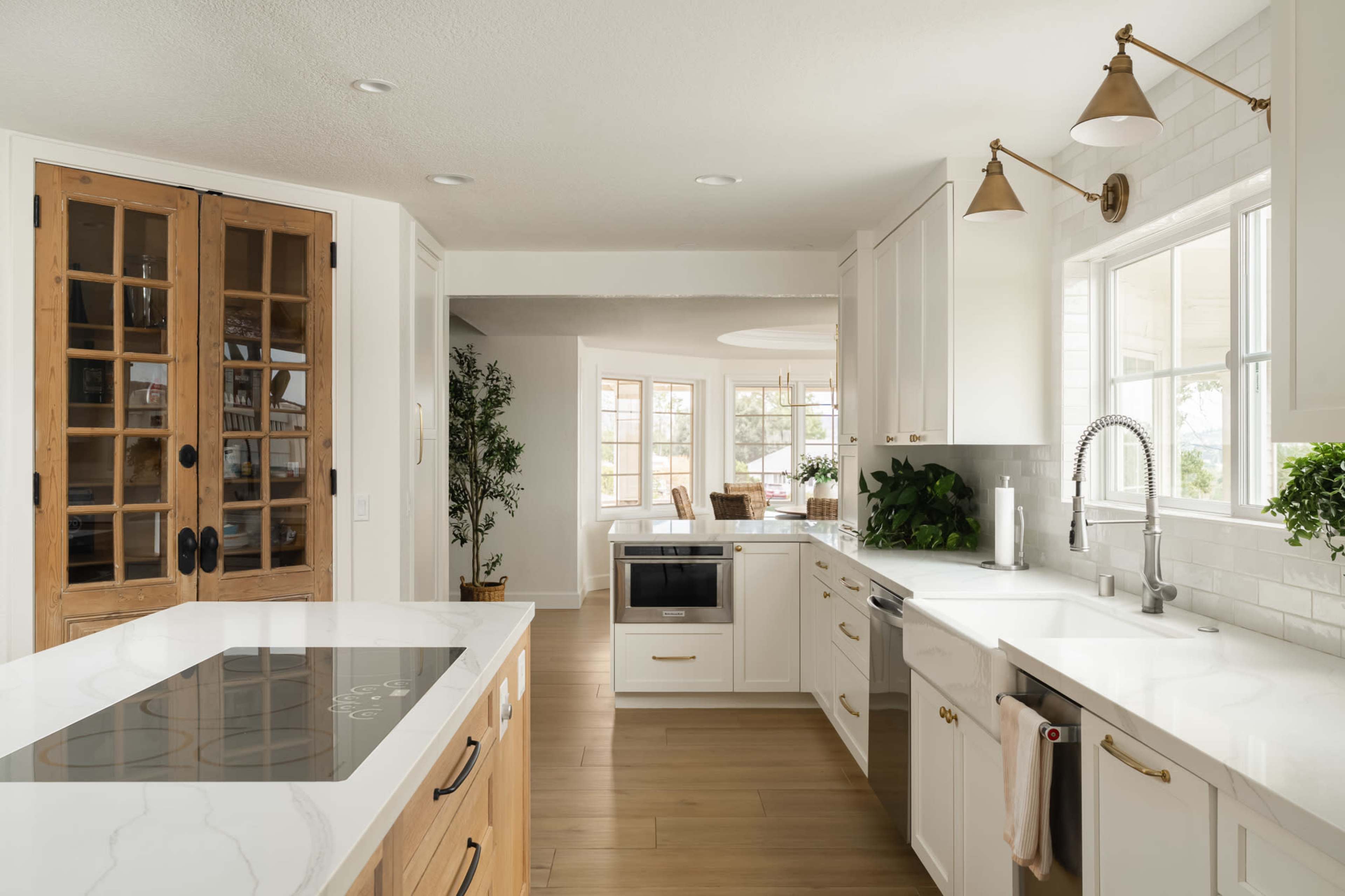 The image features a modern kitchen with white cabinets, a central island, and large windows allowing natural light to enter the space.