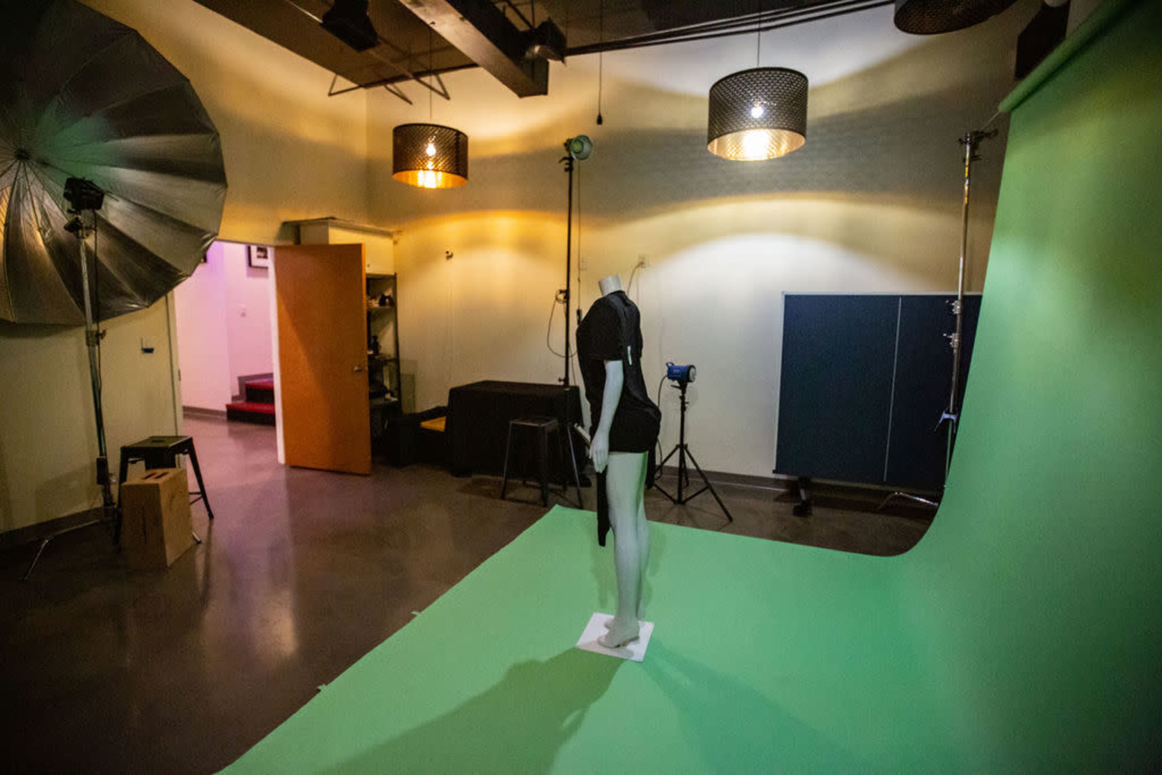 The image shows a photography studio with a green backdrop, a mannequin dressed in black, and various lighting equipment.