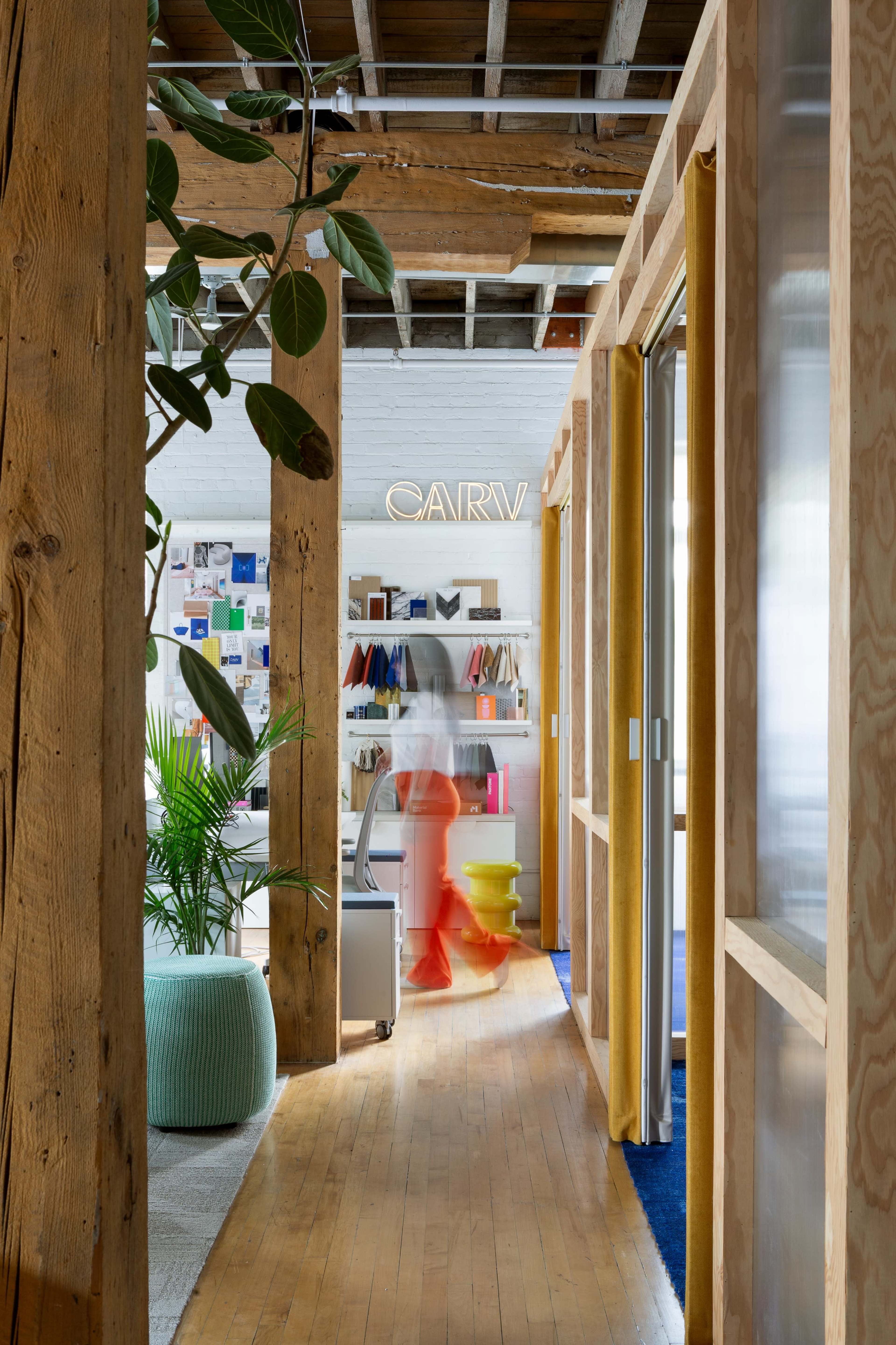 A bright office space features wooden beams, colorful decor, and a person walking past workstations.
