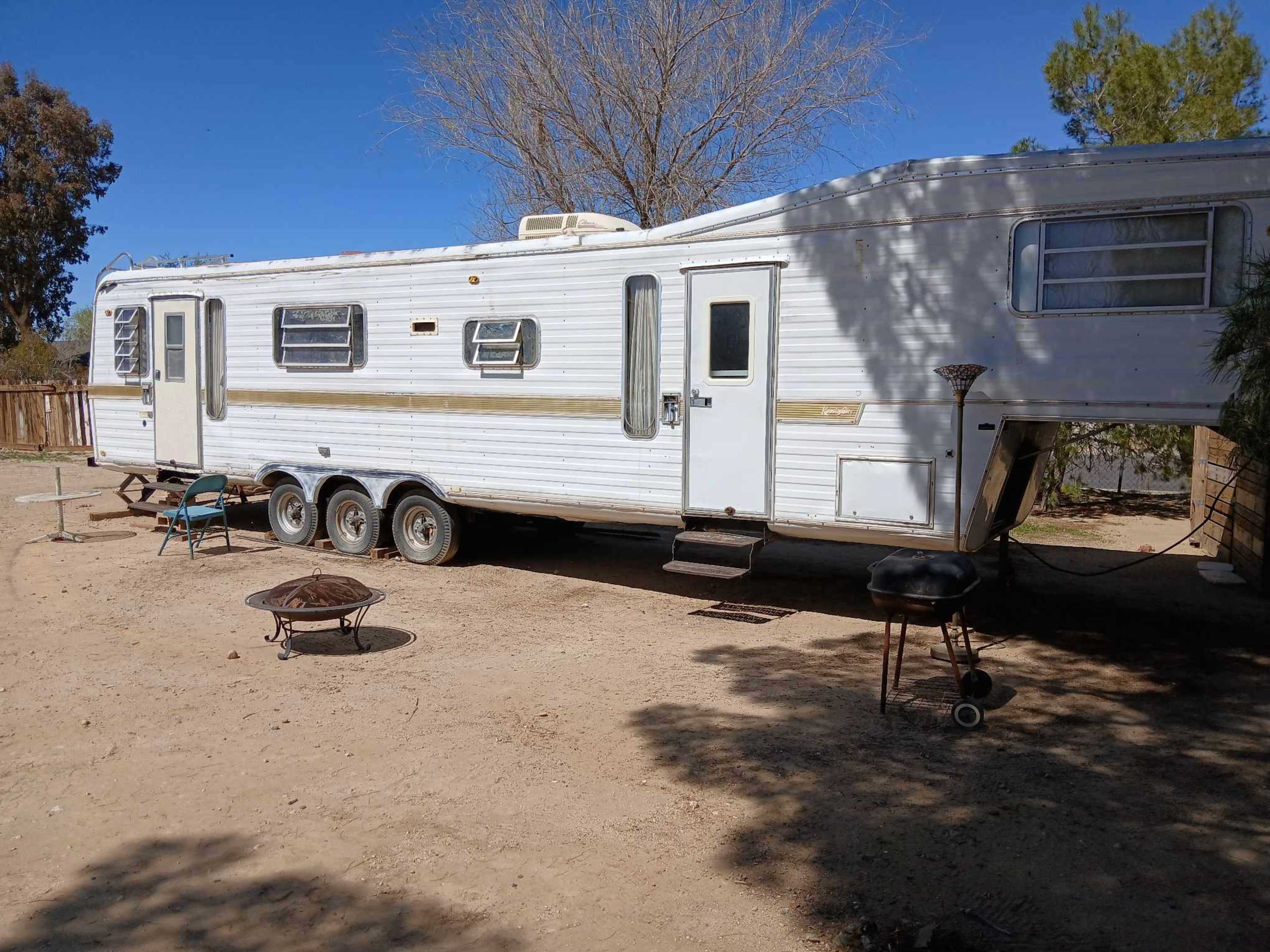 A white travel trailer is parked on a dirt lot with a small fire pit and grill nearby.