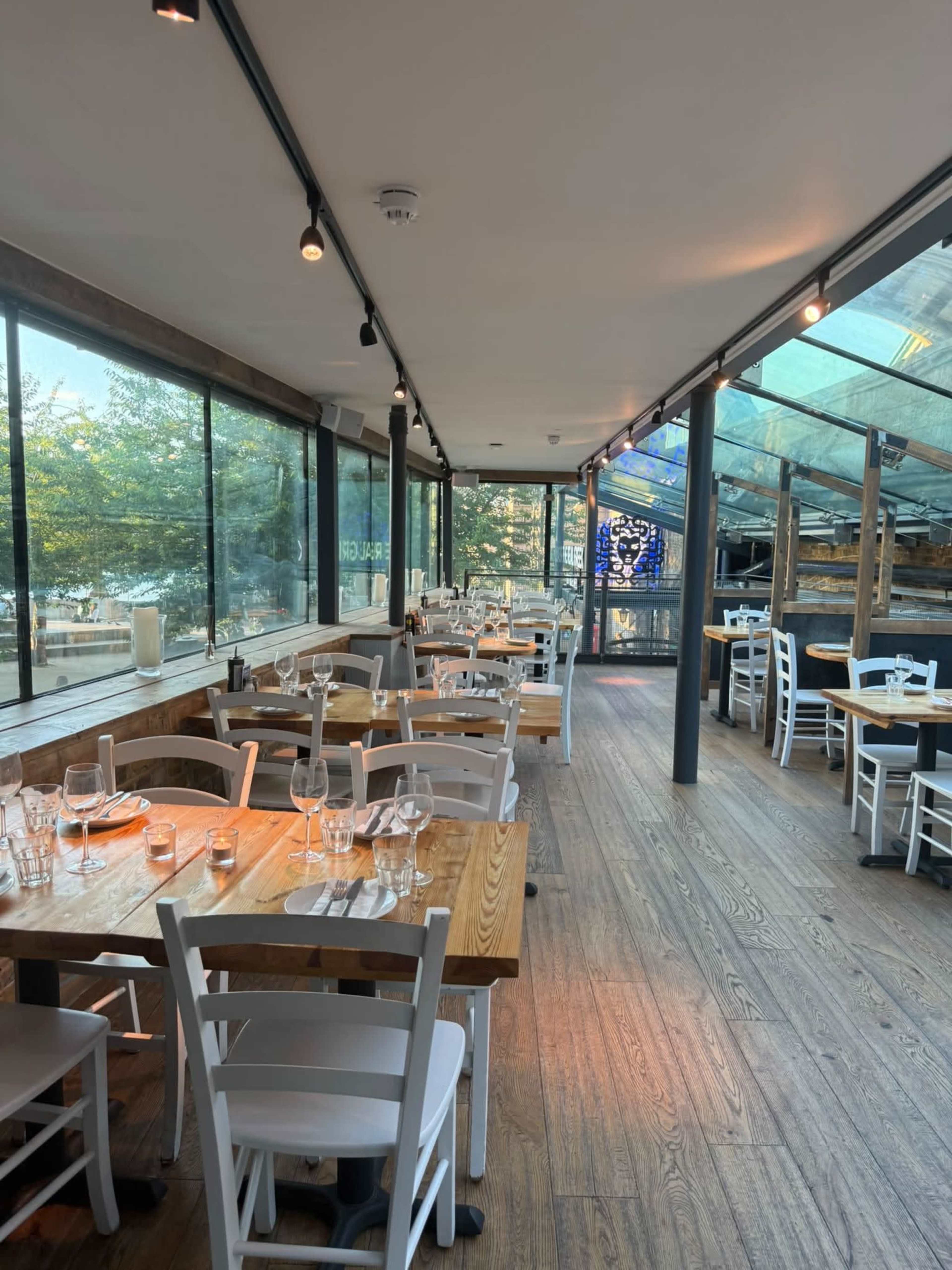 The image shows a spacious restaurant interior with wooden tables and white chairs, surrounded by large glass windows and a modern design.