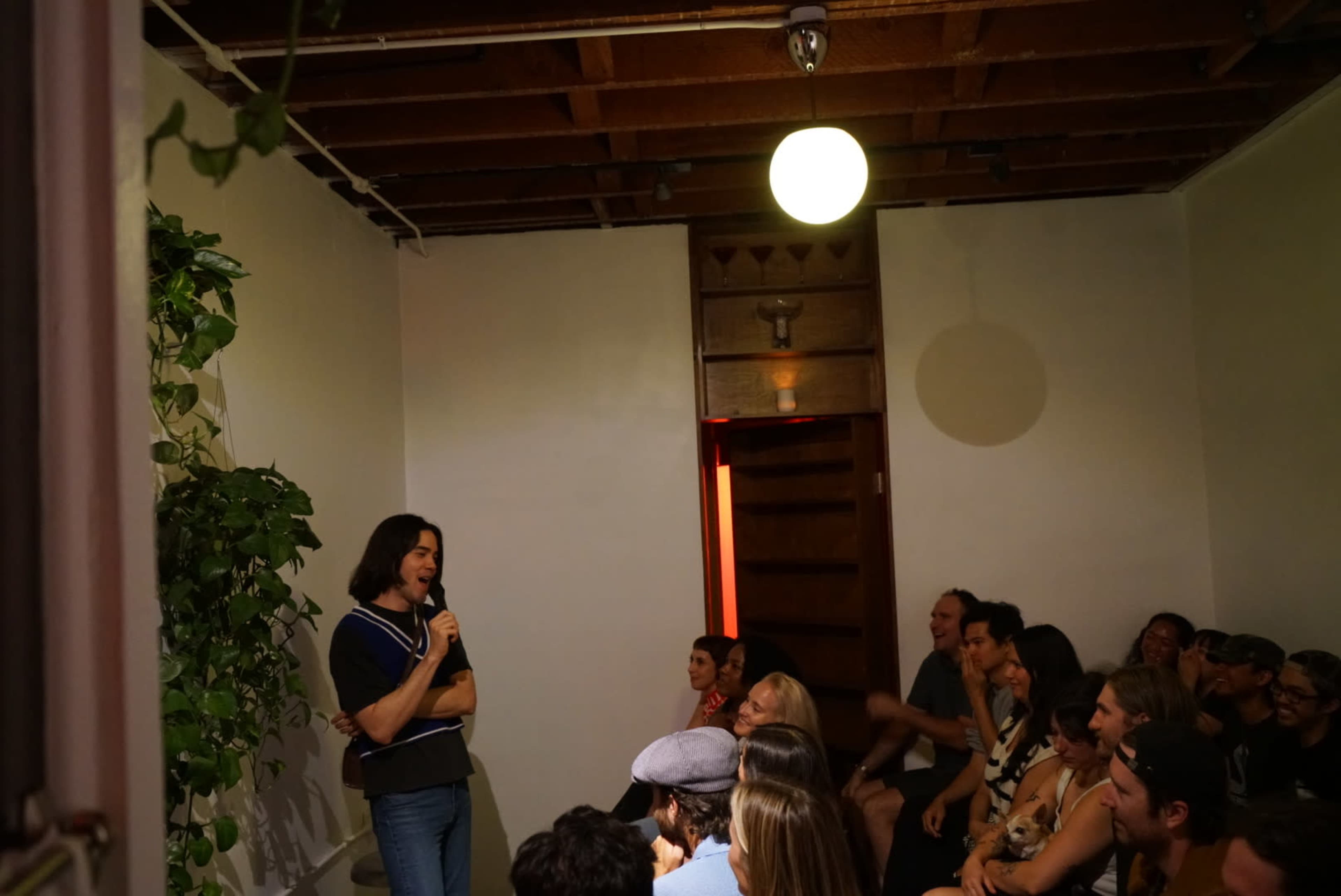 A speaker stands at a microphone in a small room filled with an audience seated on the floor and against the walls.