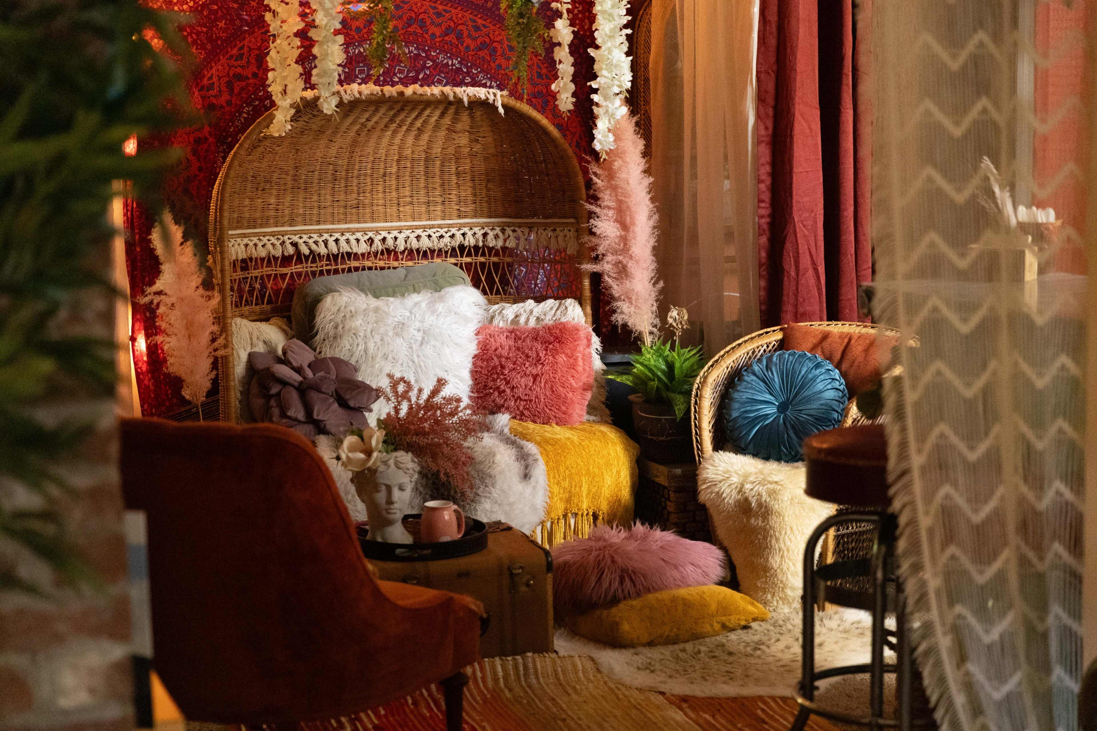 A cozy seating area features a woven rattan chair surrounded by colorful pillows, plants, and decorative fabrics.