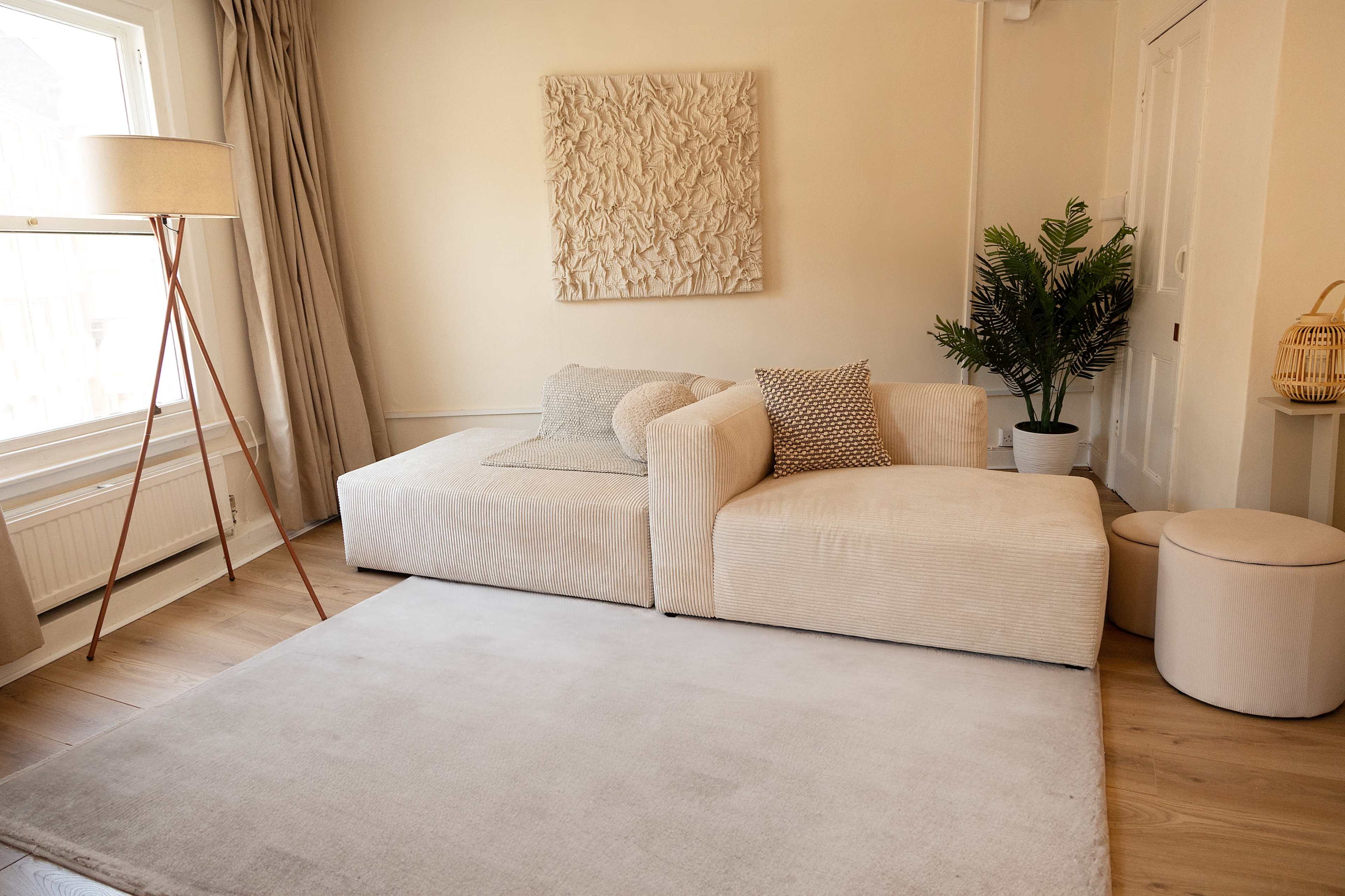 A modern, light-filled living room featuring a beige modular sofa arrangement, a tall lamp, a potted plant, and a round ottoman on a light rug.