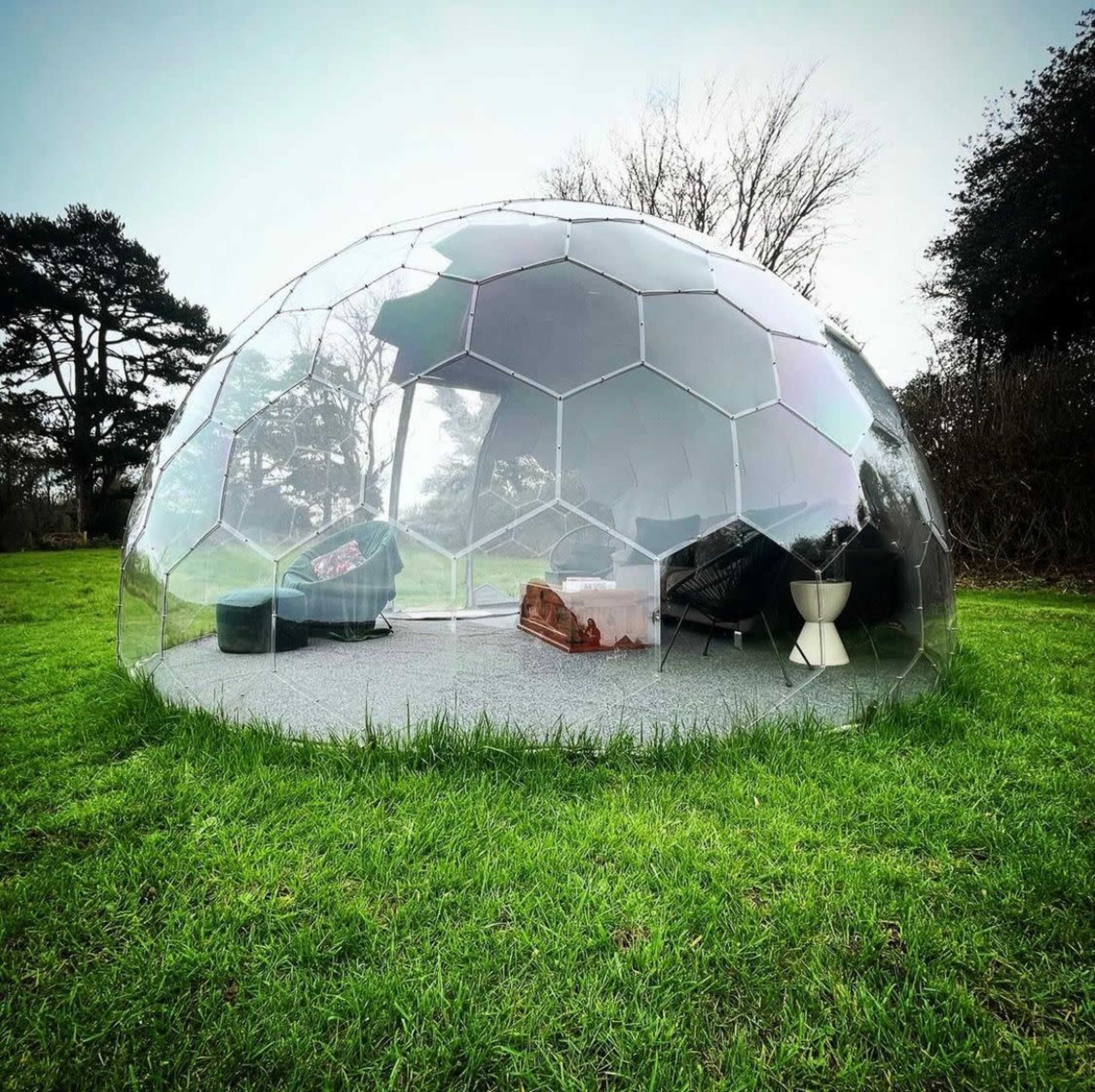 A transparent, hexagonal dome structure situated on a grassy area, furnished with a couch, table, and a chair inside.