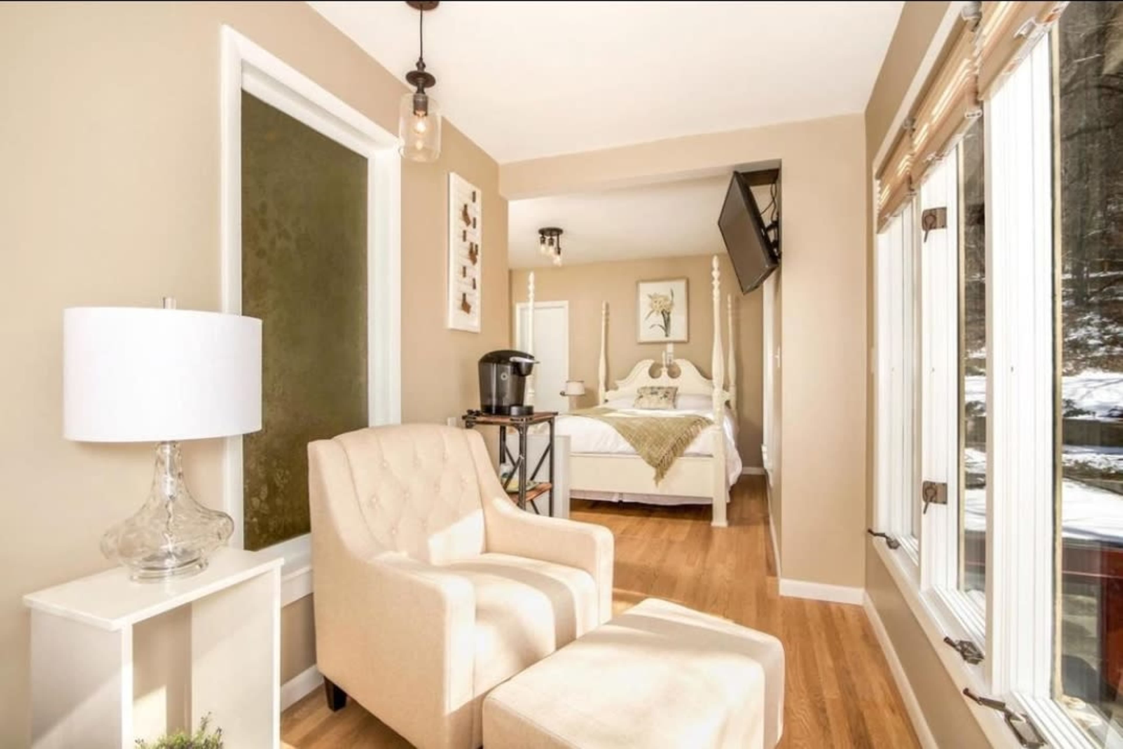 A light-colored armchair and side table are positioned in a cozy room that leads to a bedroom with a bed and a mounted TV.