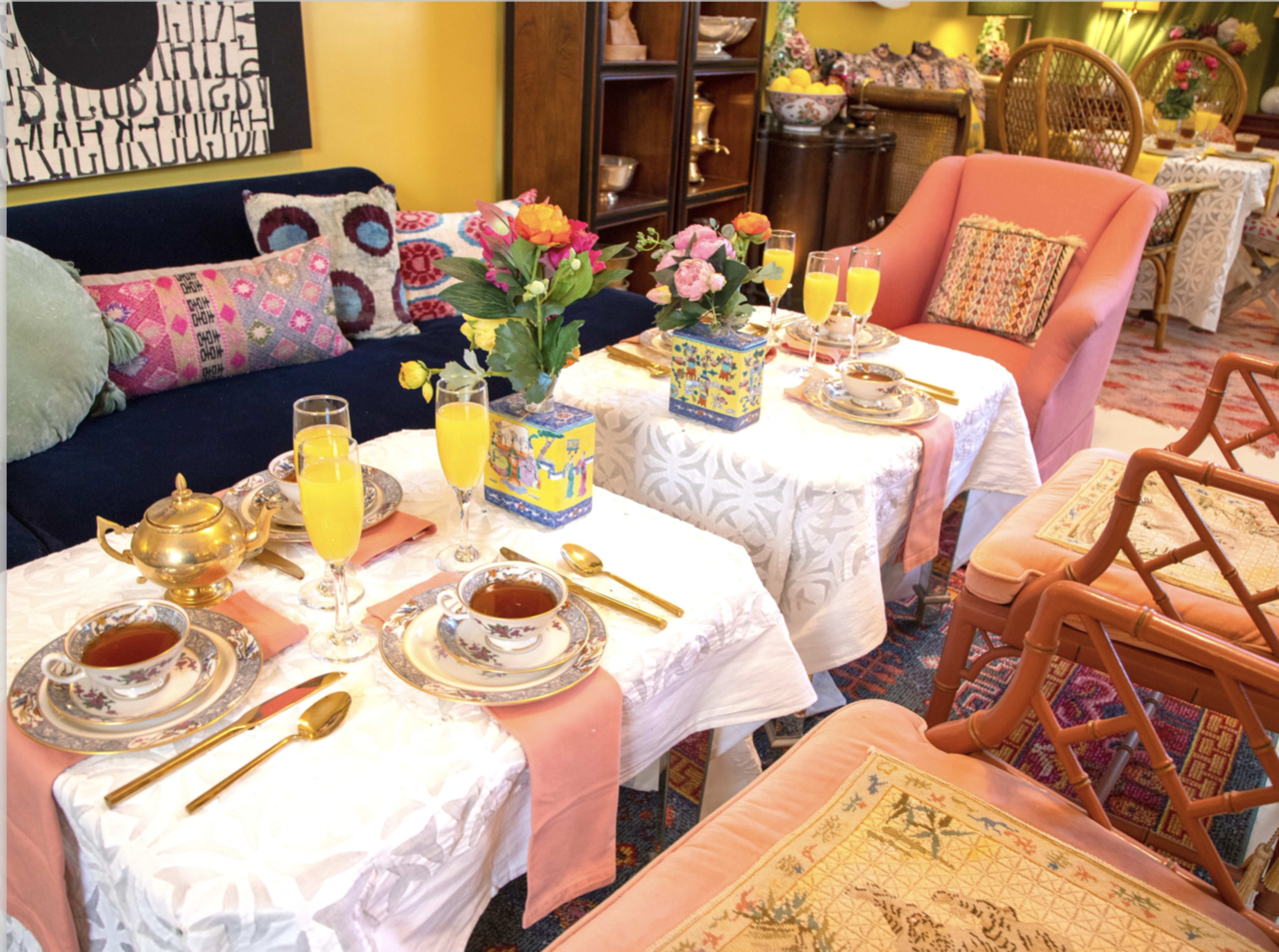 The scene features a cozy gathering area with two tables set for tea, adorned with floral arrangements, teacups, and glasses of orange juice, surrounded by colorful furniture and decor.