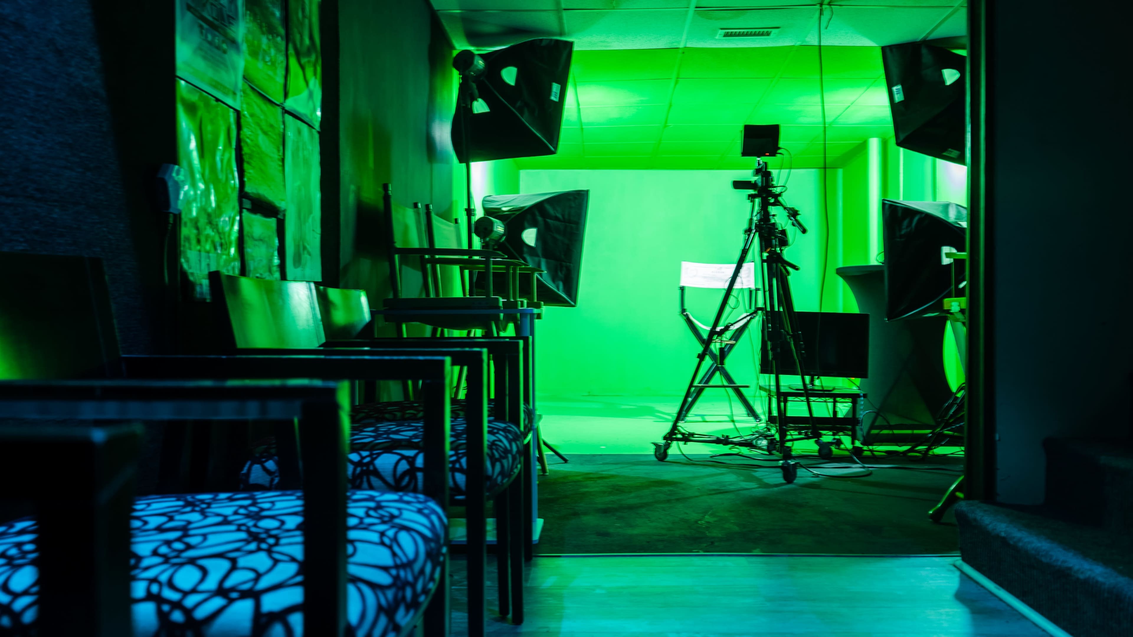 A green-lit studio space with a camera setup, lighting equipment, and chairs along the side.