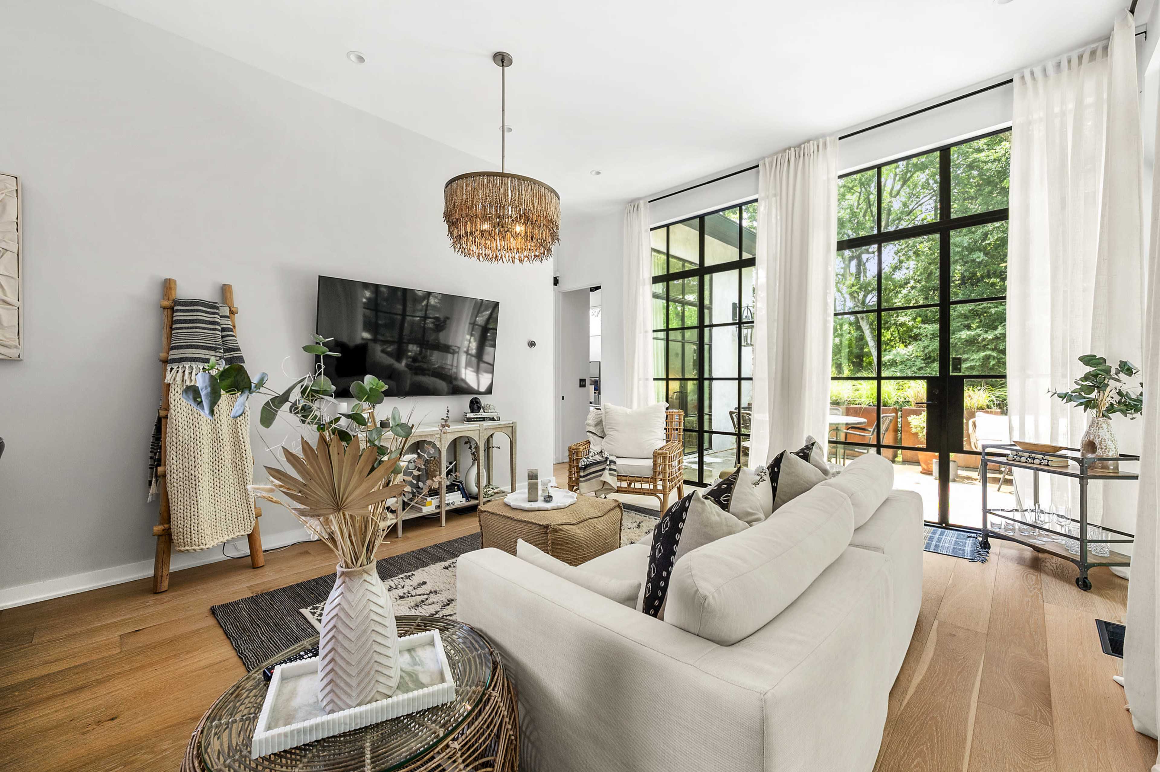 A modern living room features a light sofa, a large television mounted on the wall, and access to a patio through glass doors surrounded by greenery.