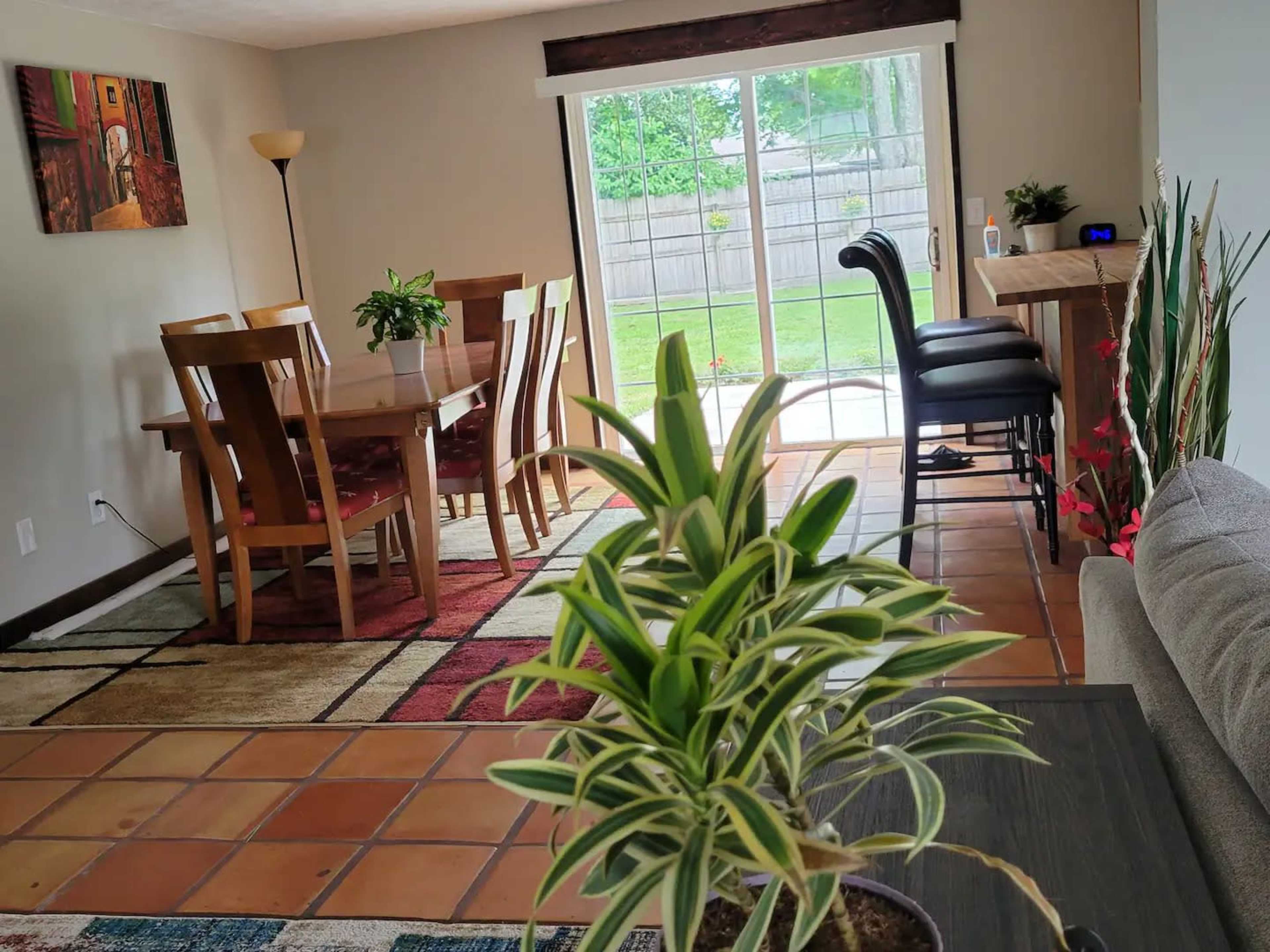 A living space with a dining table and chairs, a bar with stools, and a view of a backyard through sliding glass doors.
