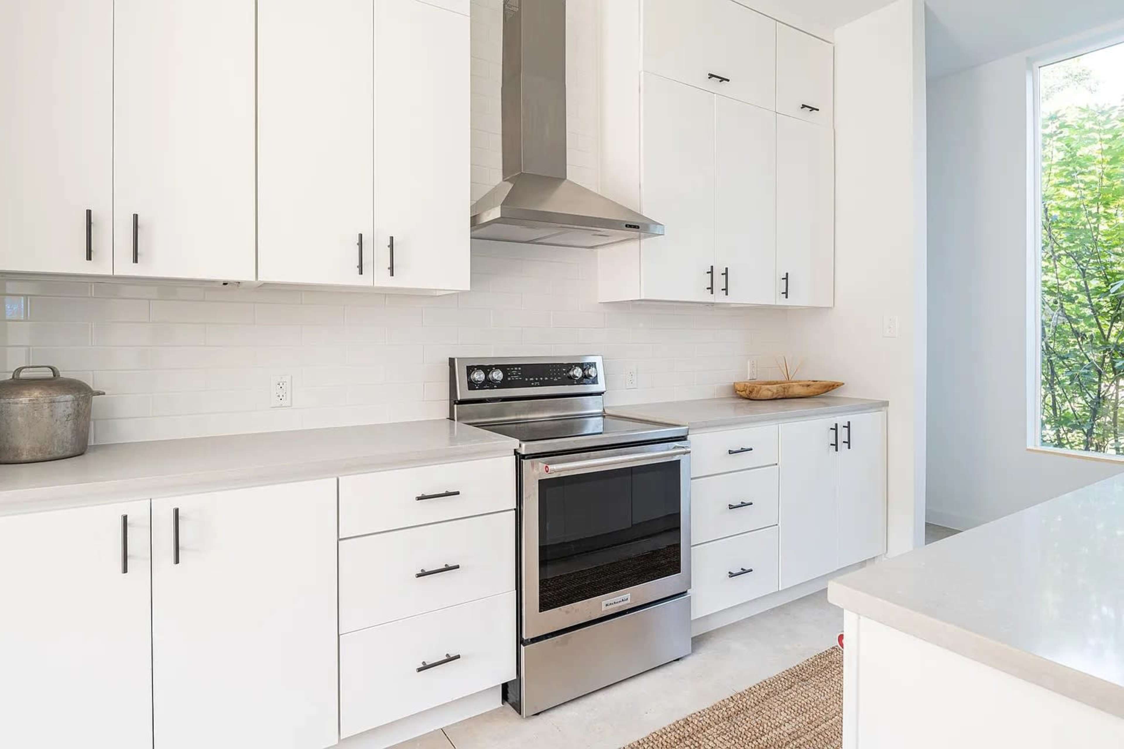 A modern kitchen features white cabinetry, a stainless steel oven, and a clean countertop with natural light streaming in through a window.