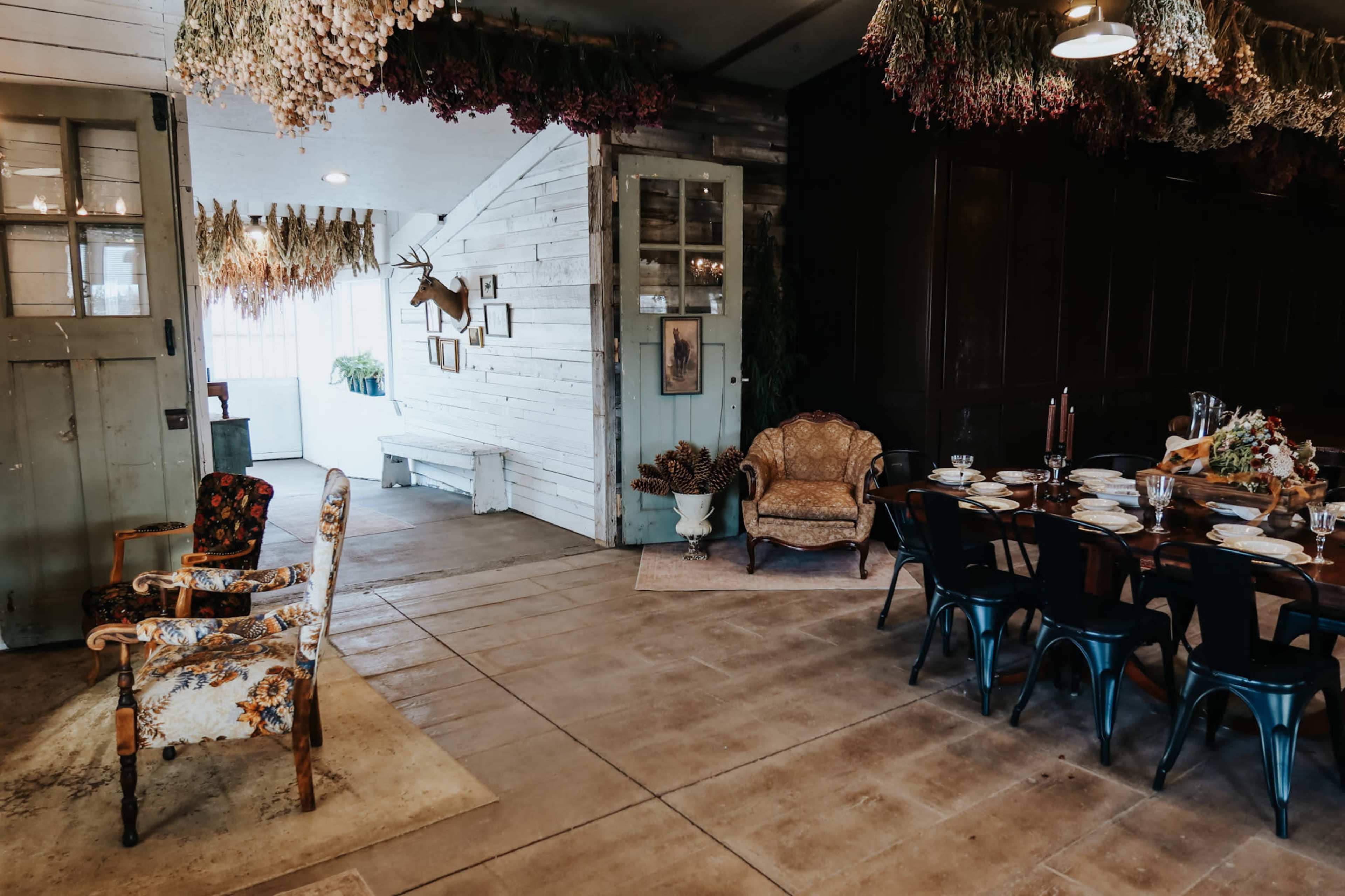 The scene features a rustic dining area with a large wooden table set for a meal, surrounded by eclectic chairs, and adorned with dried flowers hanging from the ceiling.
