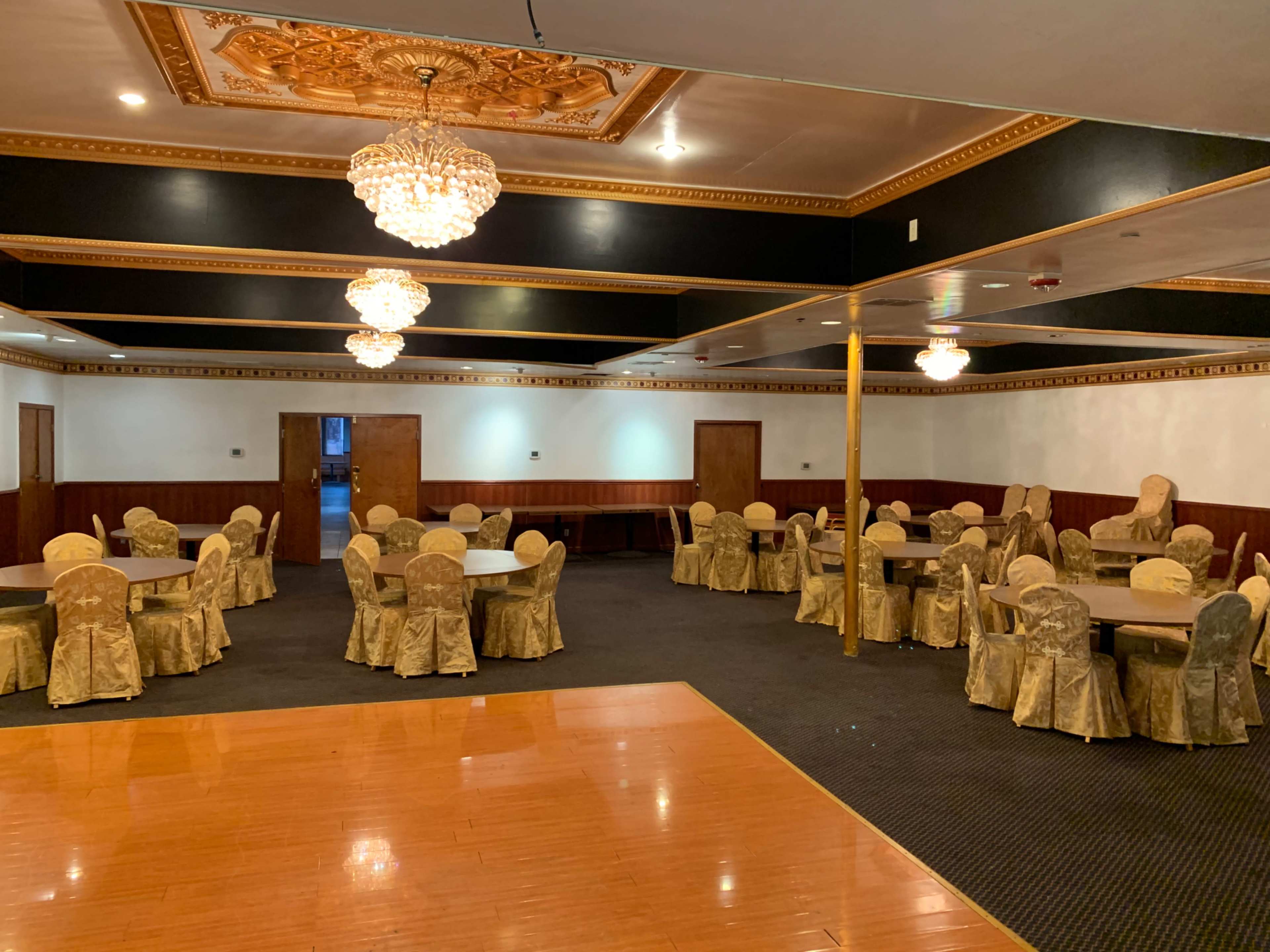 A spacious banquet hall with round tables covered in gold tablecloths, surrounded by chairs, and adorned with chandeliers hanging from the ceiling.