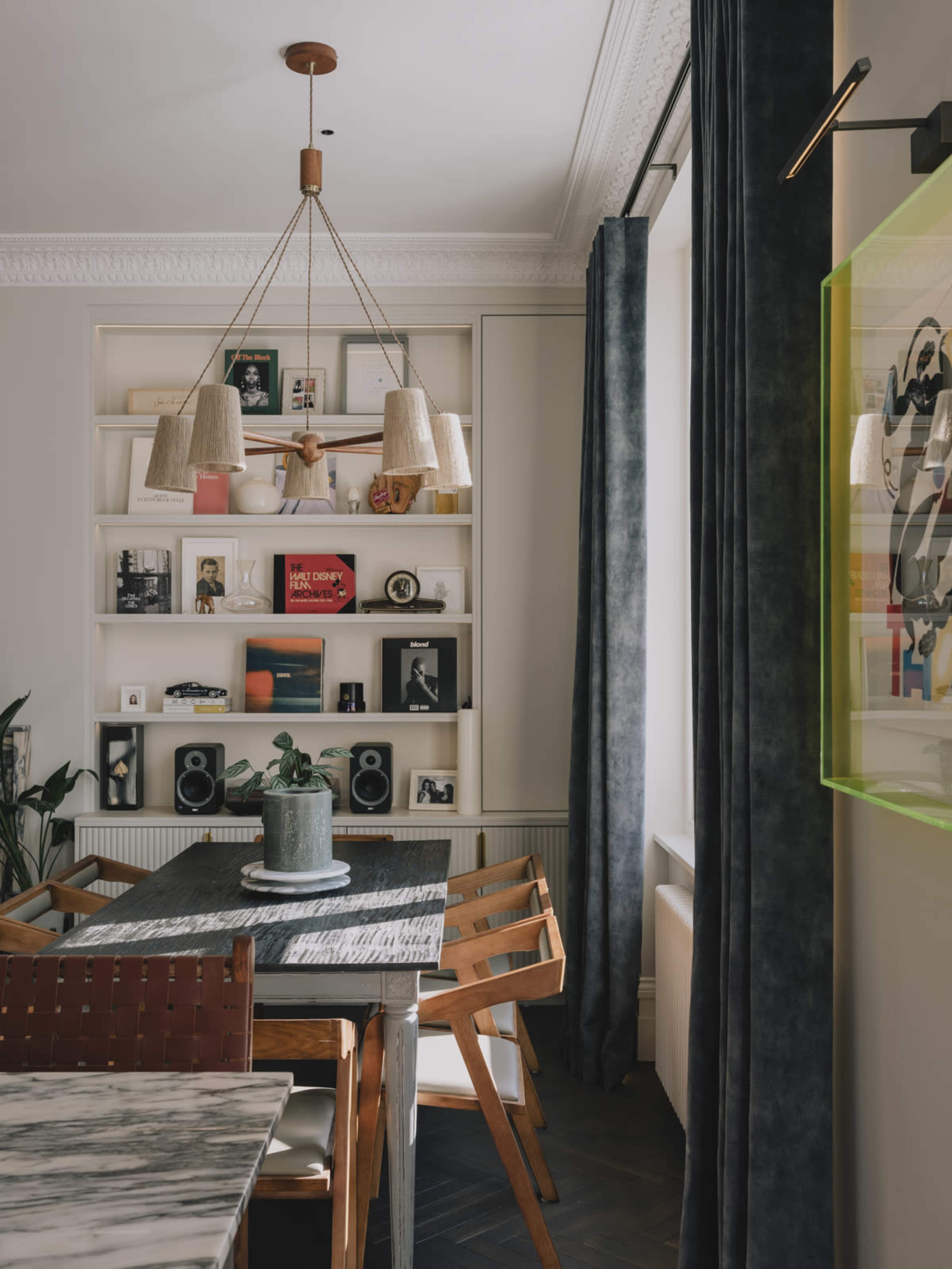 The image shows a modern dining area with a wooden table, surrounded by chairs, a pendant lamp above, and a bookshelf filled with framed photos and decor.