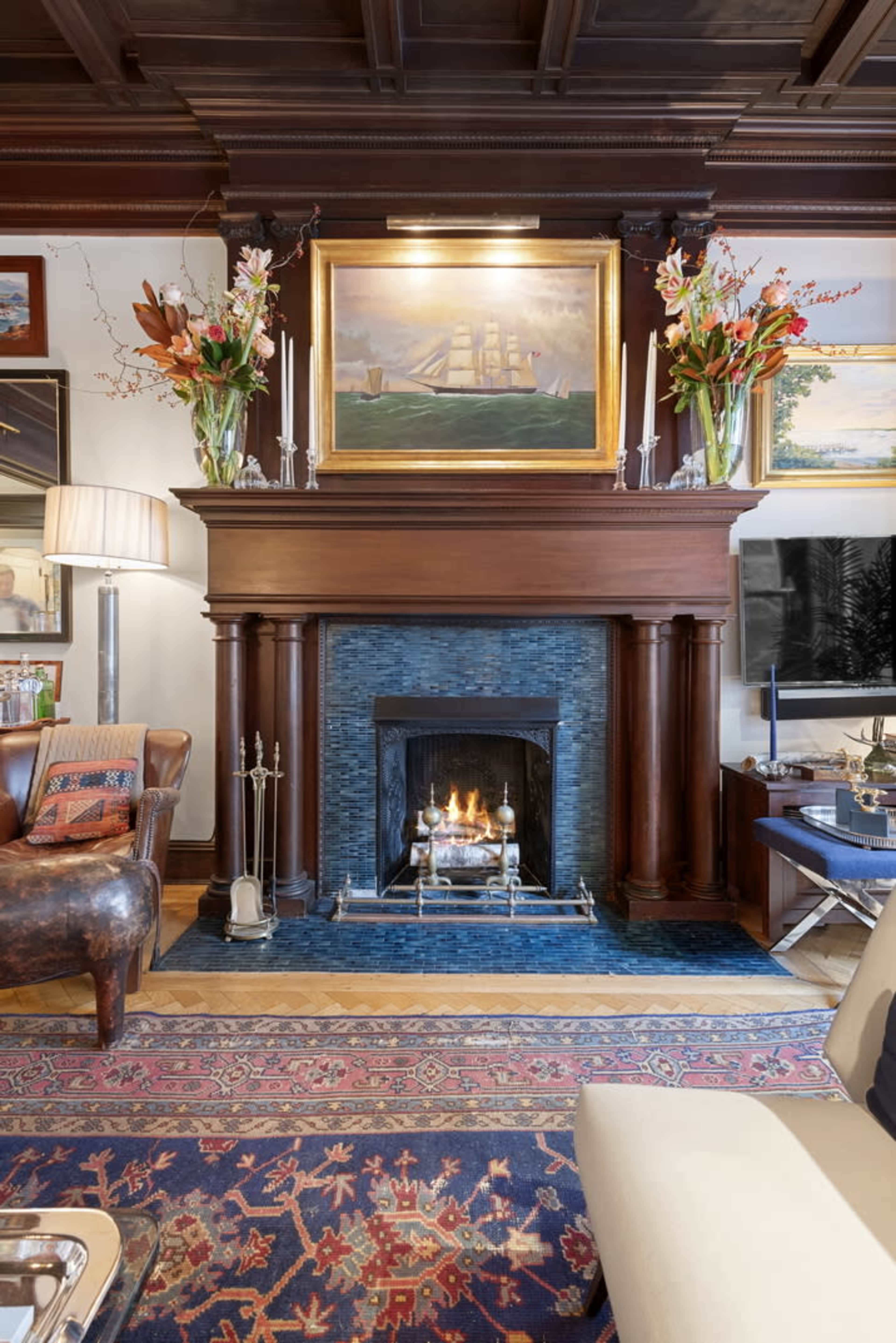 The image shows a living room featuring a fireplace with a framed painting above it, flanked by floral arrangements, a comfortable chair, and a rug on the floor.