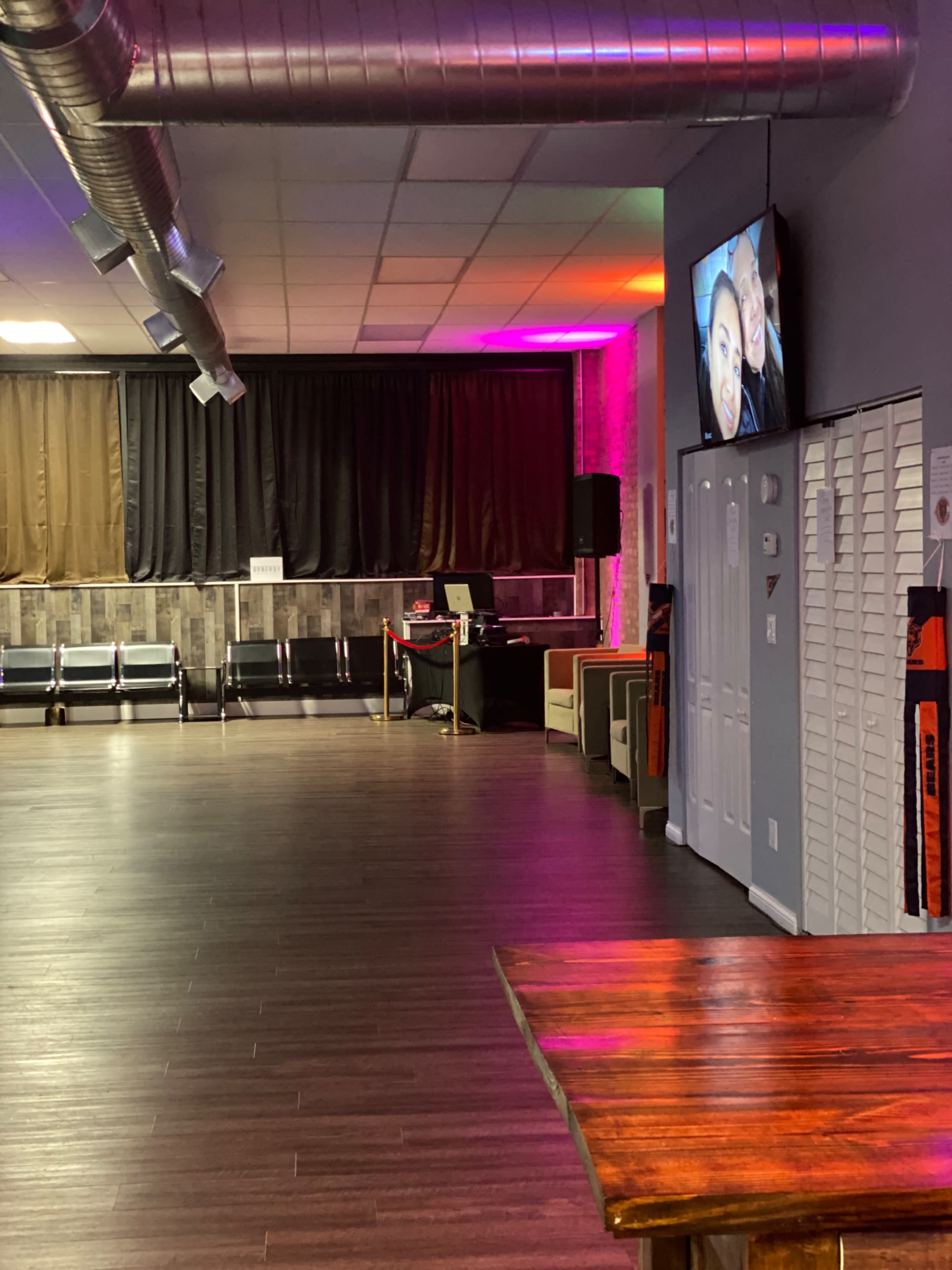 The image shows an interior space with wooden flooring, seating areas, a reception desk, and a television on the wall displaying a person's face.