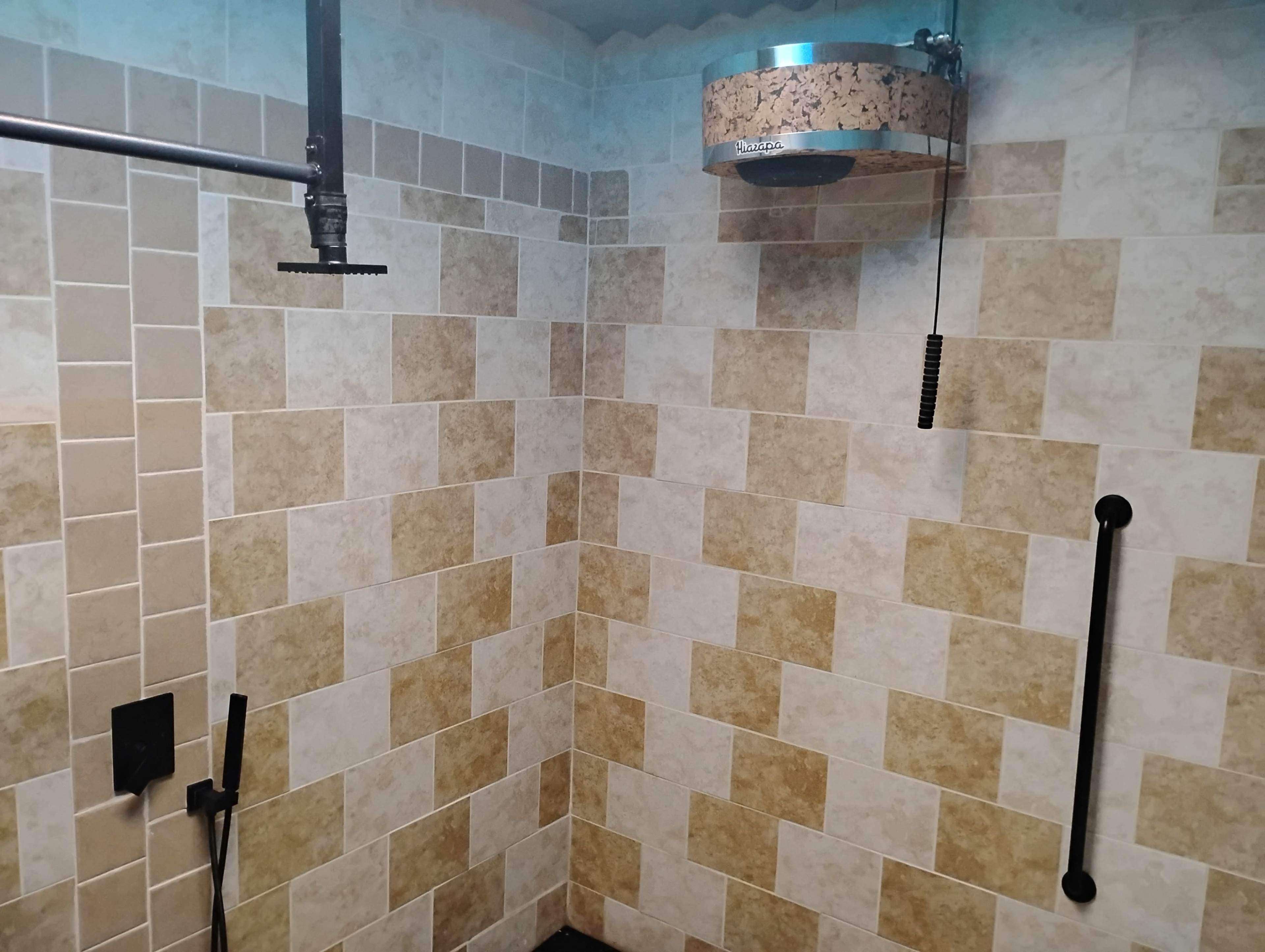 The image shows a shower area with tiled walls in a pattern of light and dark squares, featuring a rainfall showerhead and a grab bar.