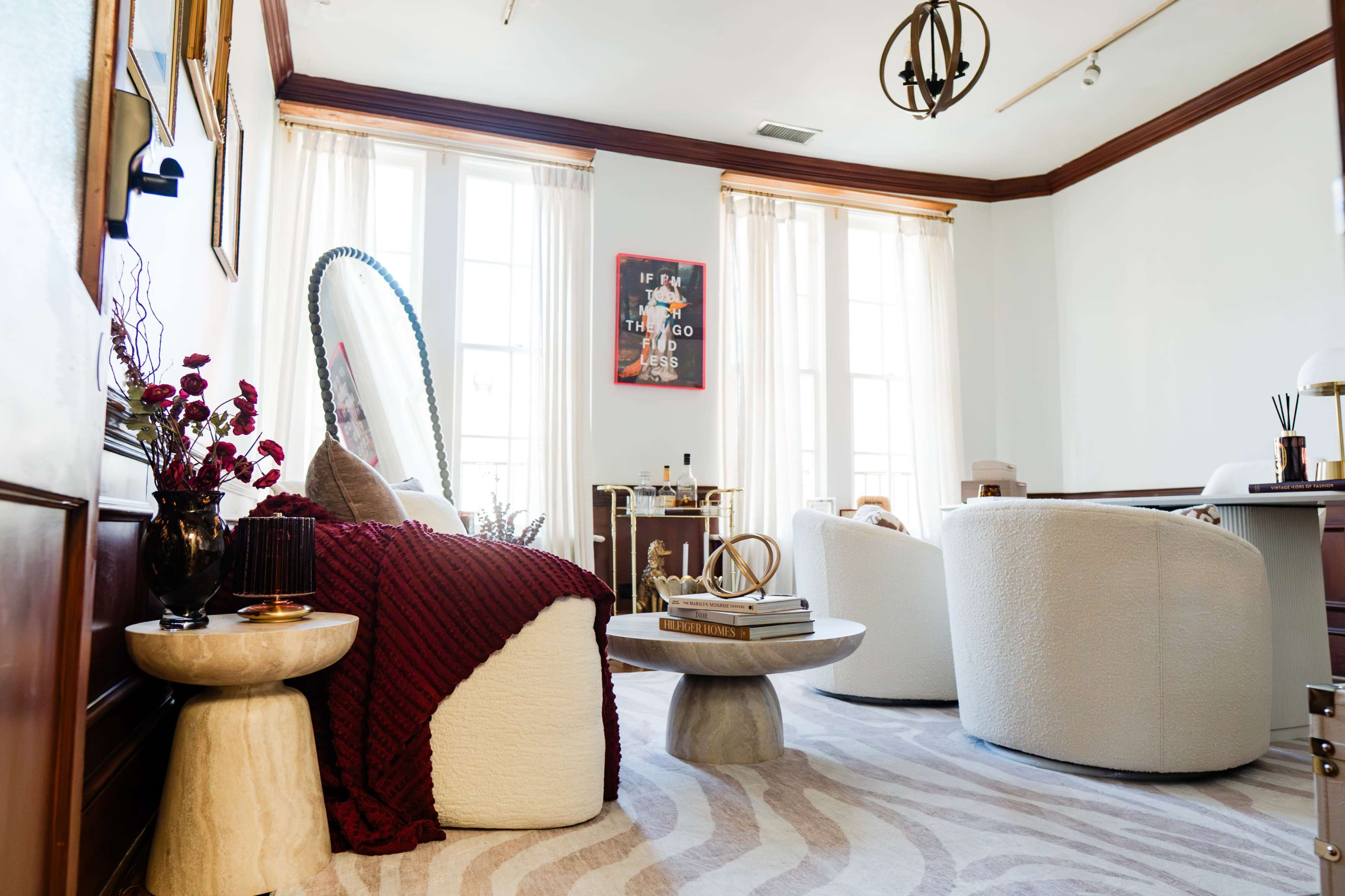 The image shows a well-lit, stylish sitting room featuring plush armchairs, a cozy throw, a round coffee table, and large windows adorned with sheer curtains.