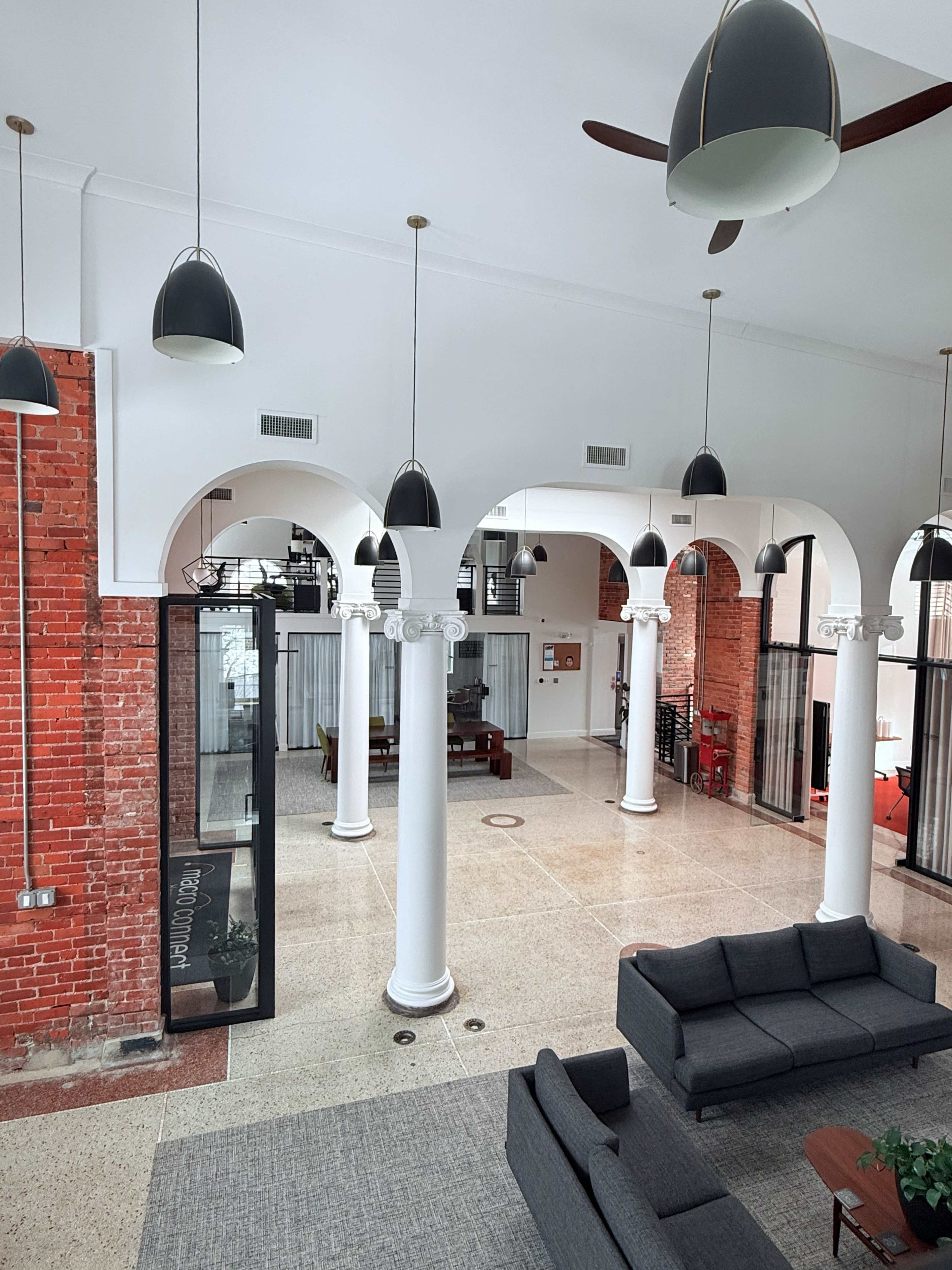 The image shows a spacious interior with tall arched columns, modern lighting fixtures, and a mix of brick and white walls, creating an open and inviting atmosphere.