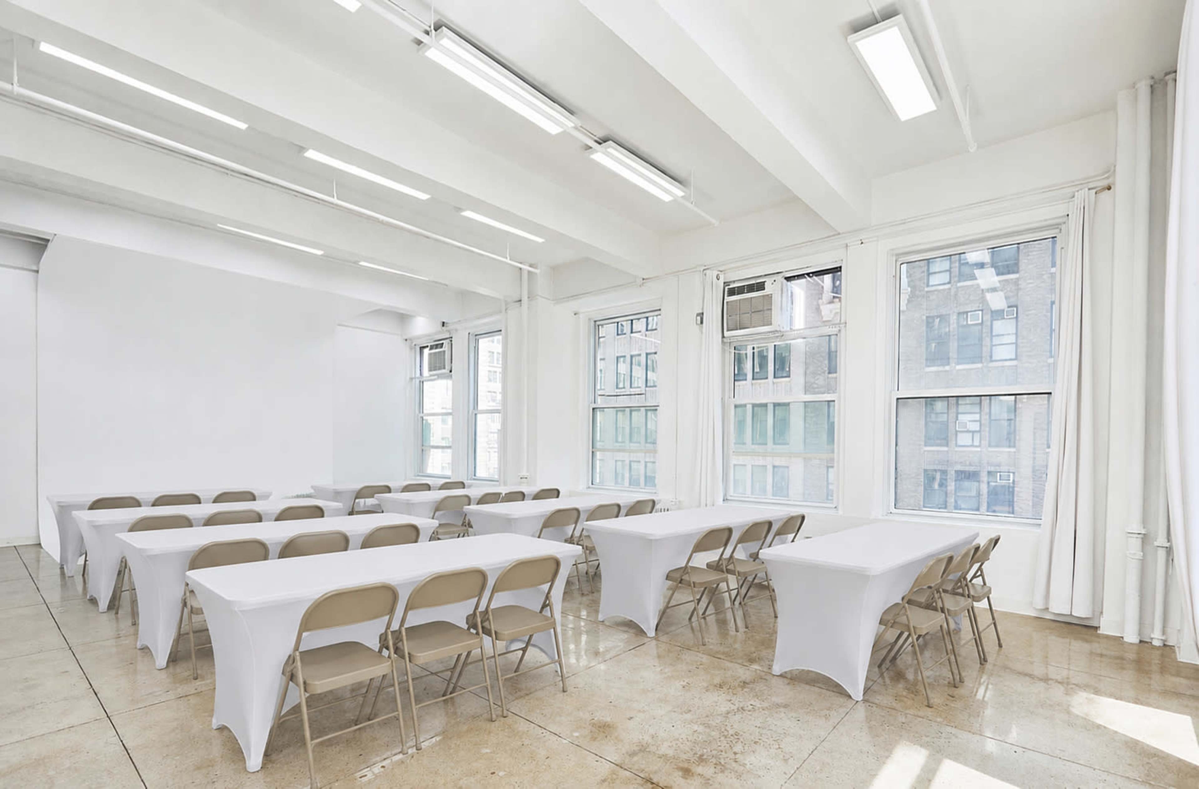 A bright, spacious room features several tables set in a U-shape, covered with white tablecloths, and surrounded by beige chairs.