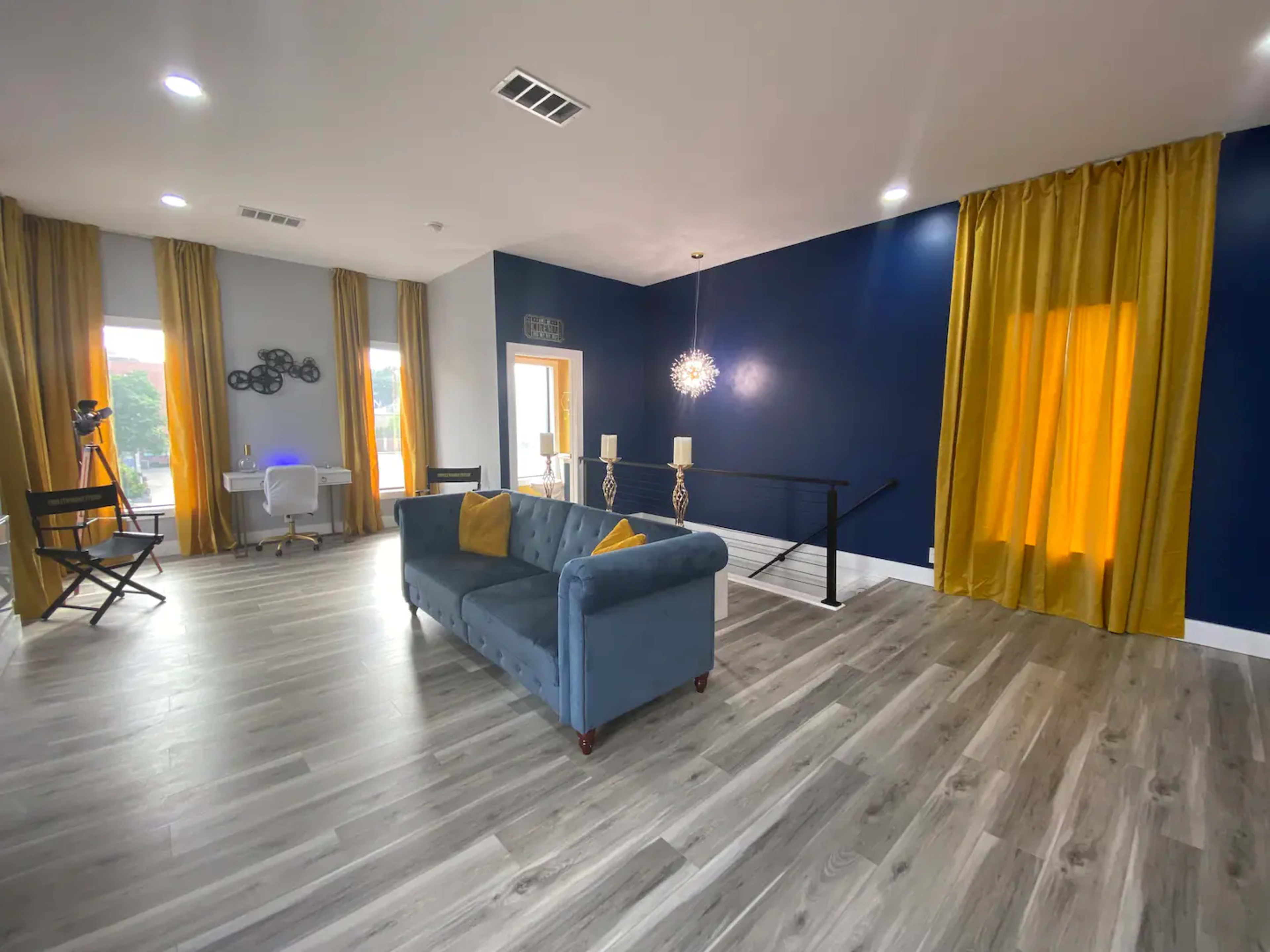 A modern living room with a gray couch, large windows with yellow curtains, and a mix of natural and artificial lighting.
